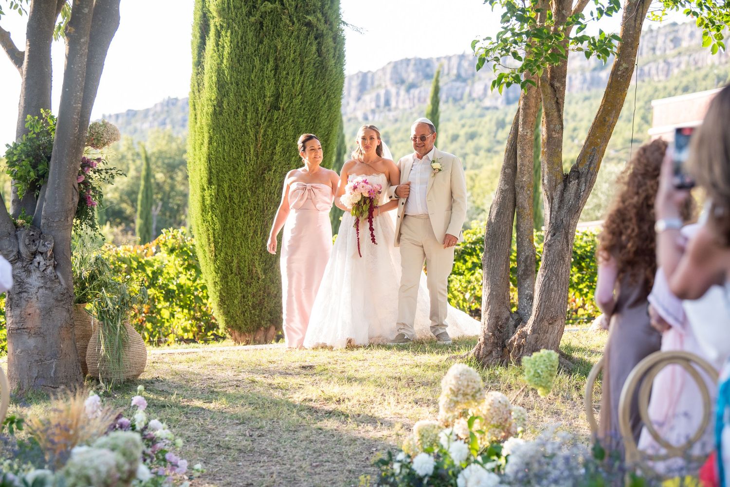 A wedding party poses together in a sunlit garden setting with trees and greenery in the background.