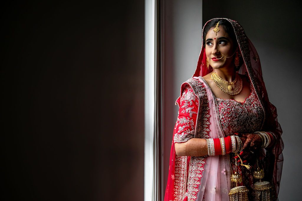 Indian bride in red lehenga and dupatta with gold jewelry poses elegantly by a window in soft natural light.