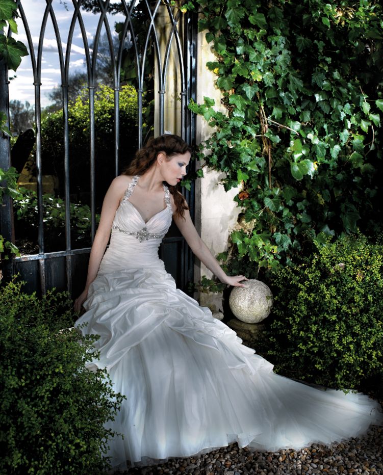 Bride in elegant white ruffled wedding gown with crystal straps poses gracefully by ivy-covered iron garden gate.