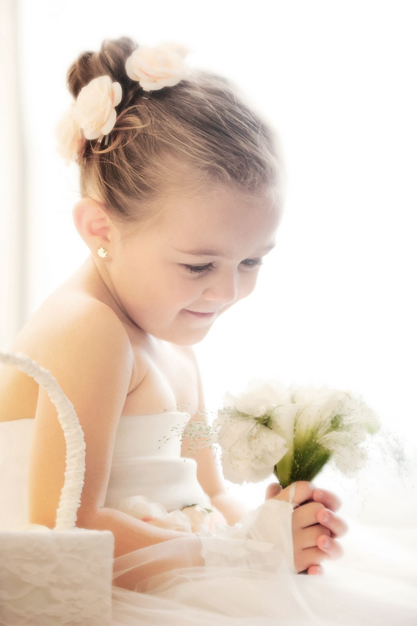 Young flower girl in white dress holding bouquet of white flowers with pink floral hair accessory.