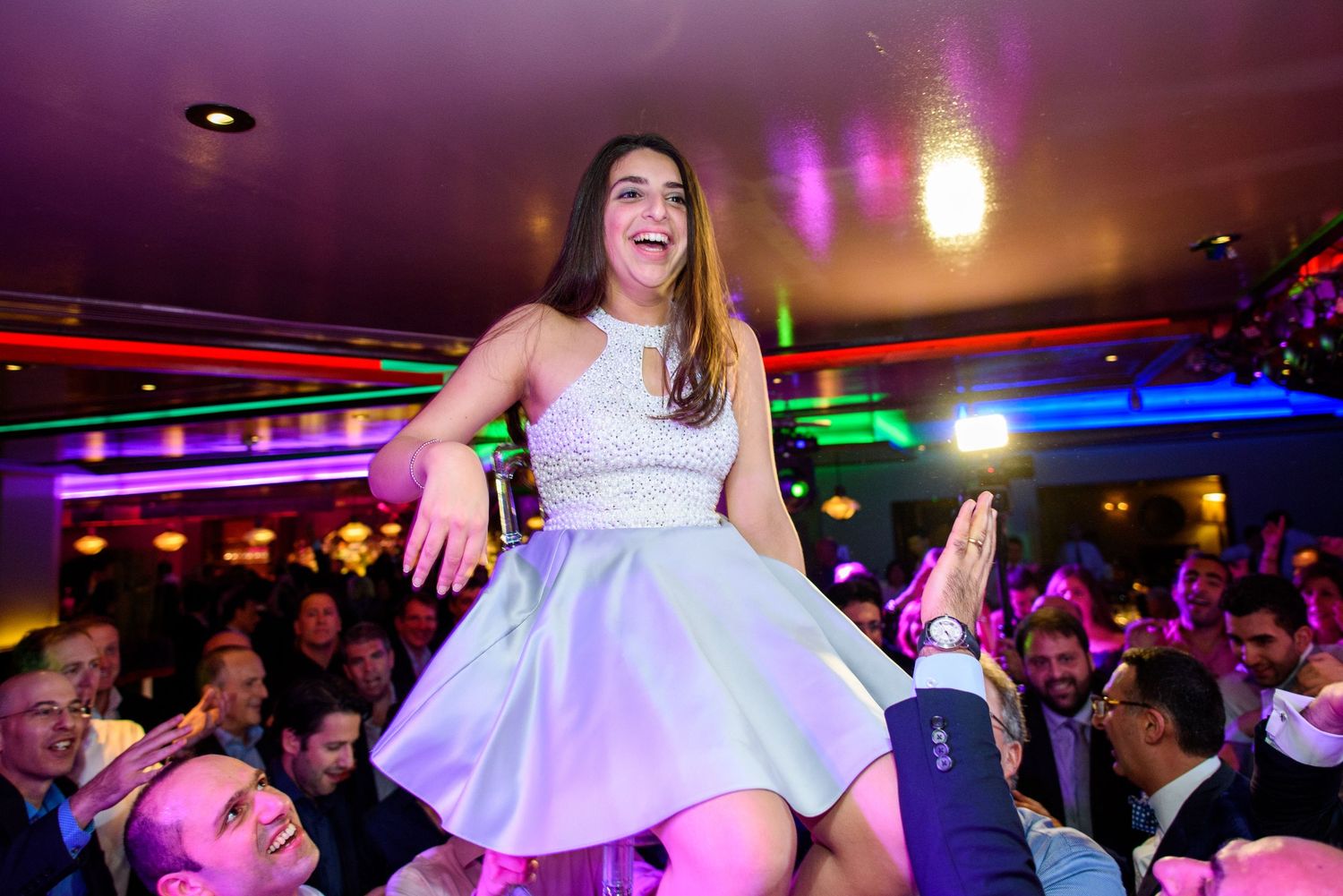 A person in a white dress dances on a raised platform at a nightclub with colorful lighting and a crowd below.