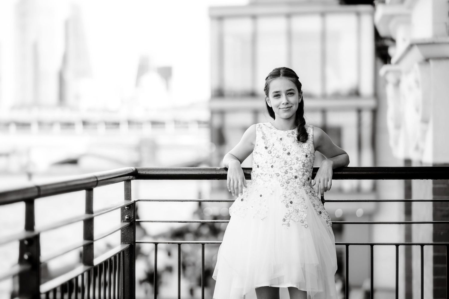 Black and white photo of someone in a white dress standing on a balcony with railing and architectural details in background.