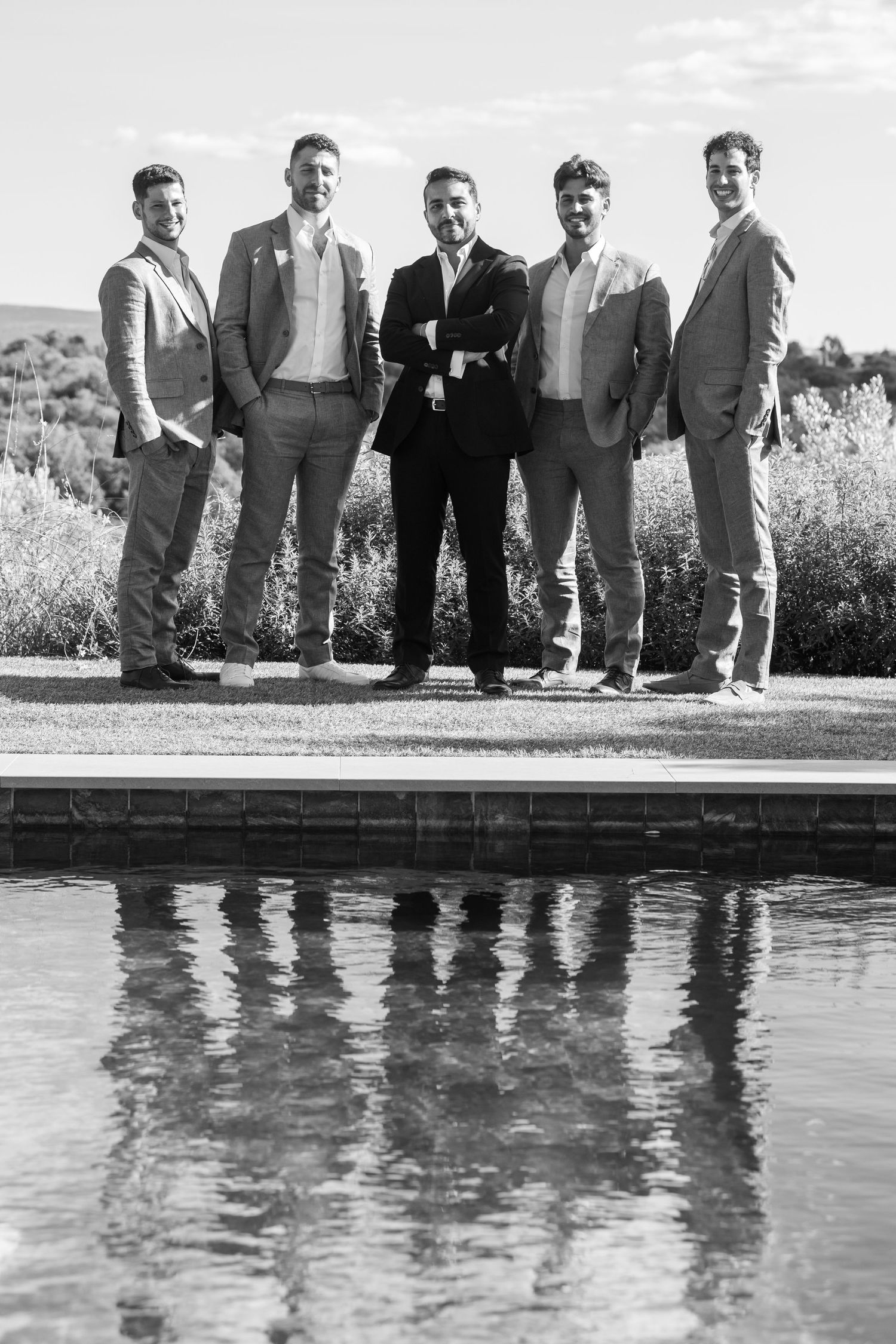 Five men in formal attire stand beside a reflective pool creating mirror images in black and white photography.