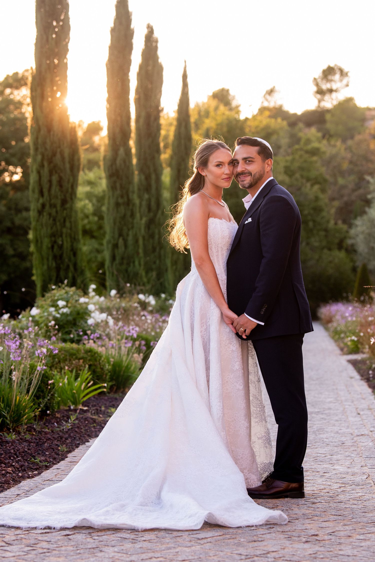 A couple poses for a romantic sunset wedding photo among cypress trees and flowering gardens.
