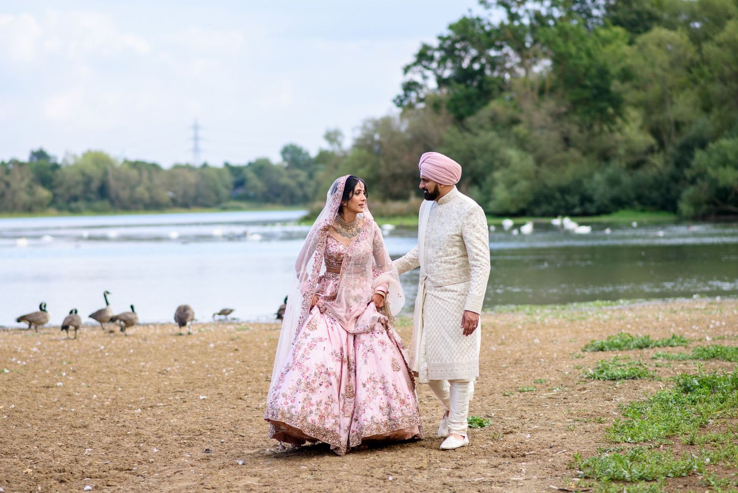 A couple in traditional attire walks along a scenic lakeside path with geese in the background.