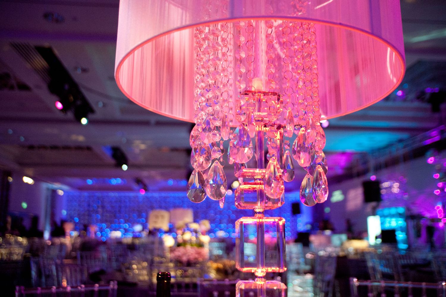Pink and purple uplighting illuminates an elegant ballroom with crystal chandeliers and luxurious table settings.