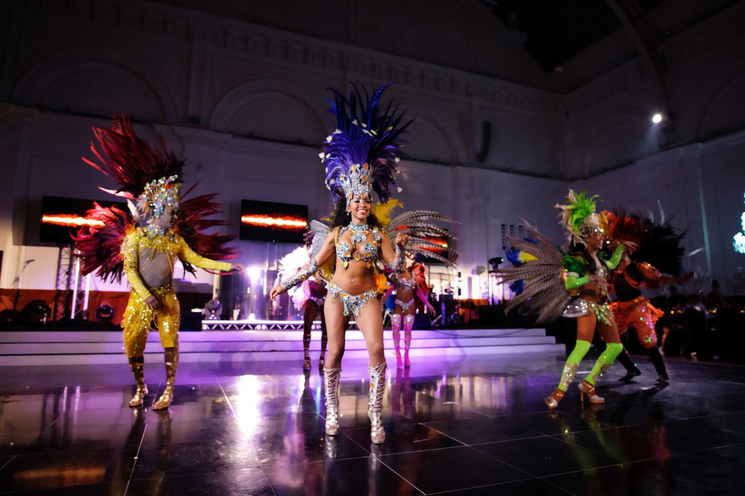 Colorfully costumed carnival dancers perform in a vibrant nighttime show on an illuminated stage.