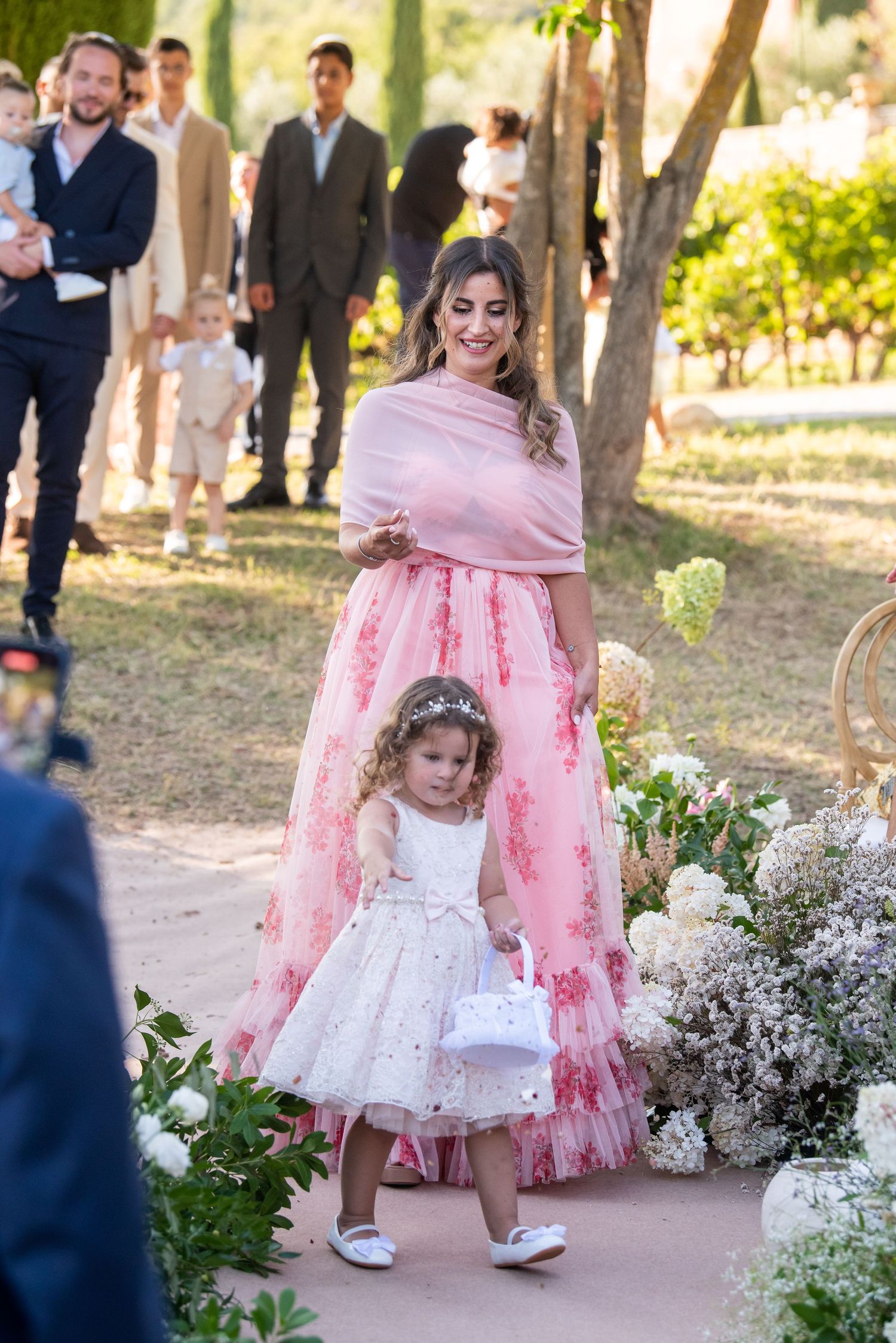 Young flower girls in pink and white dresses walk along garden path decorated with white flowers at outdoor wedding ceremony.