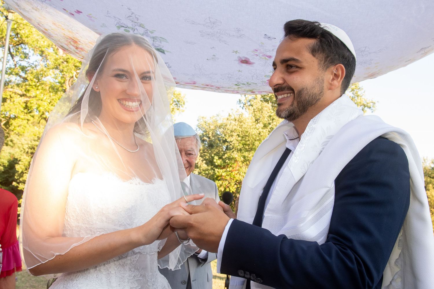 A couple exchanges rings during an outdoor wedding ceremony under a white canopy on a sunny day.