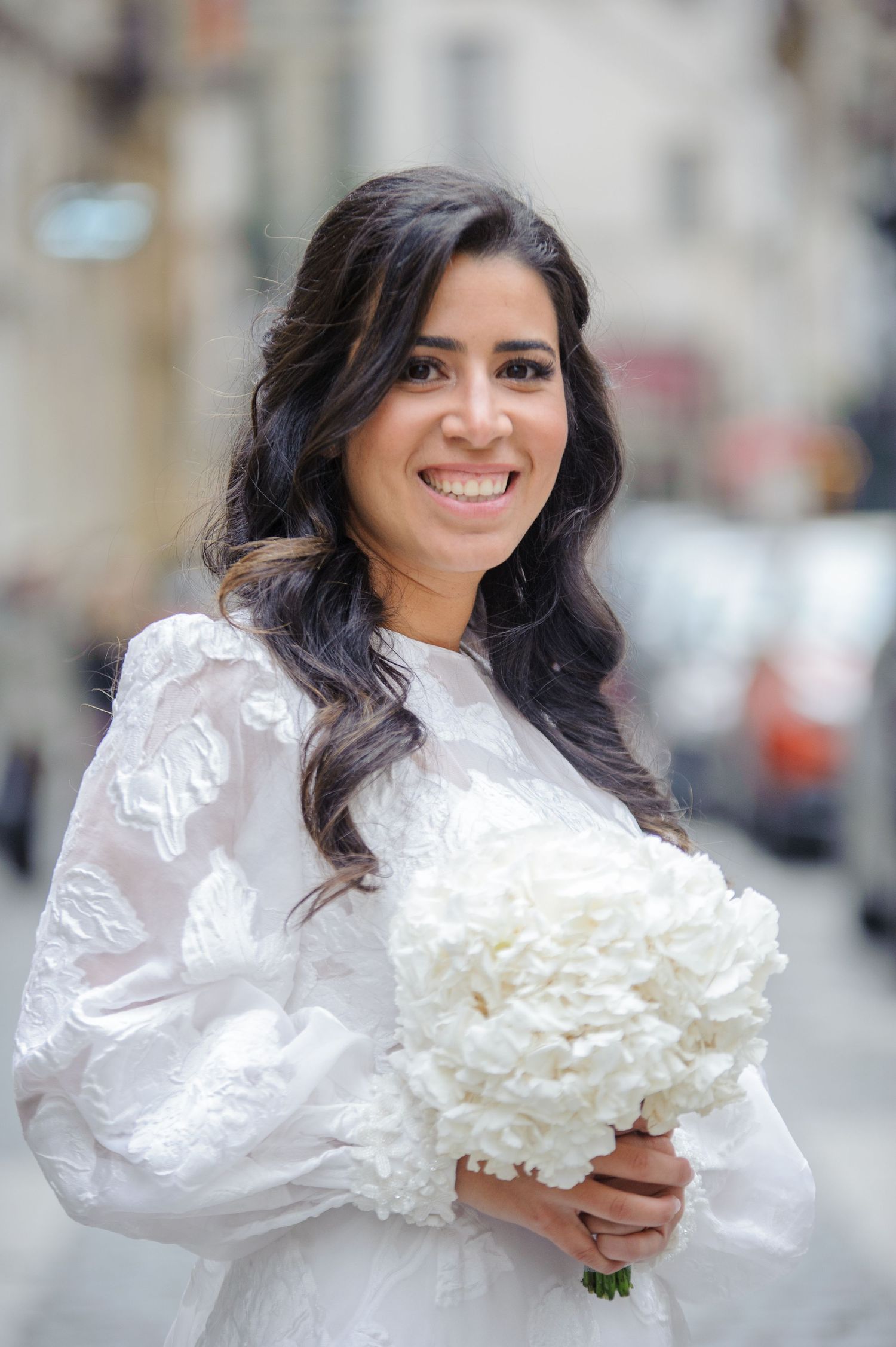 Bride in white lace dress holding white bouquet smiles while posing on city street.