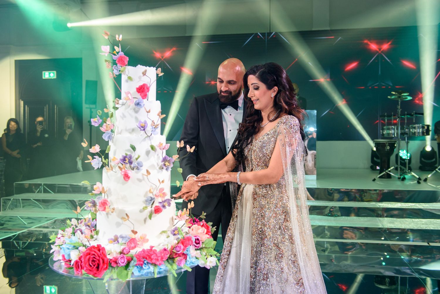 Bride and groom cutting a tall floral white wedding cake on a mirrored dance floor with colorful stage lighting.