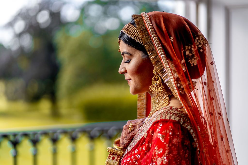 Indian bride in red bridal lehenga and gold jewelry smiling in profile, wearing traditional dupatta outdoors.
