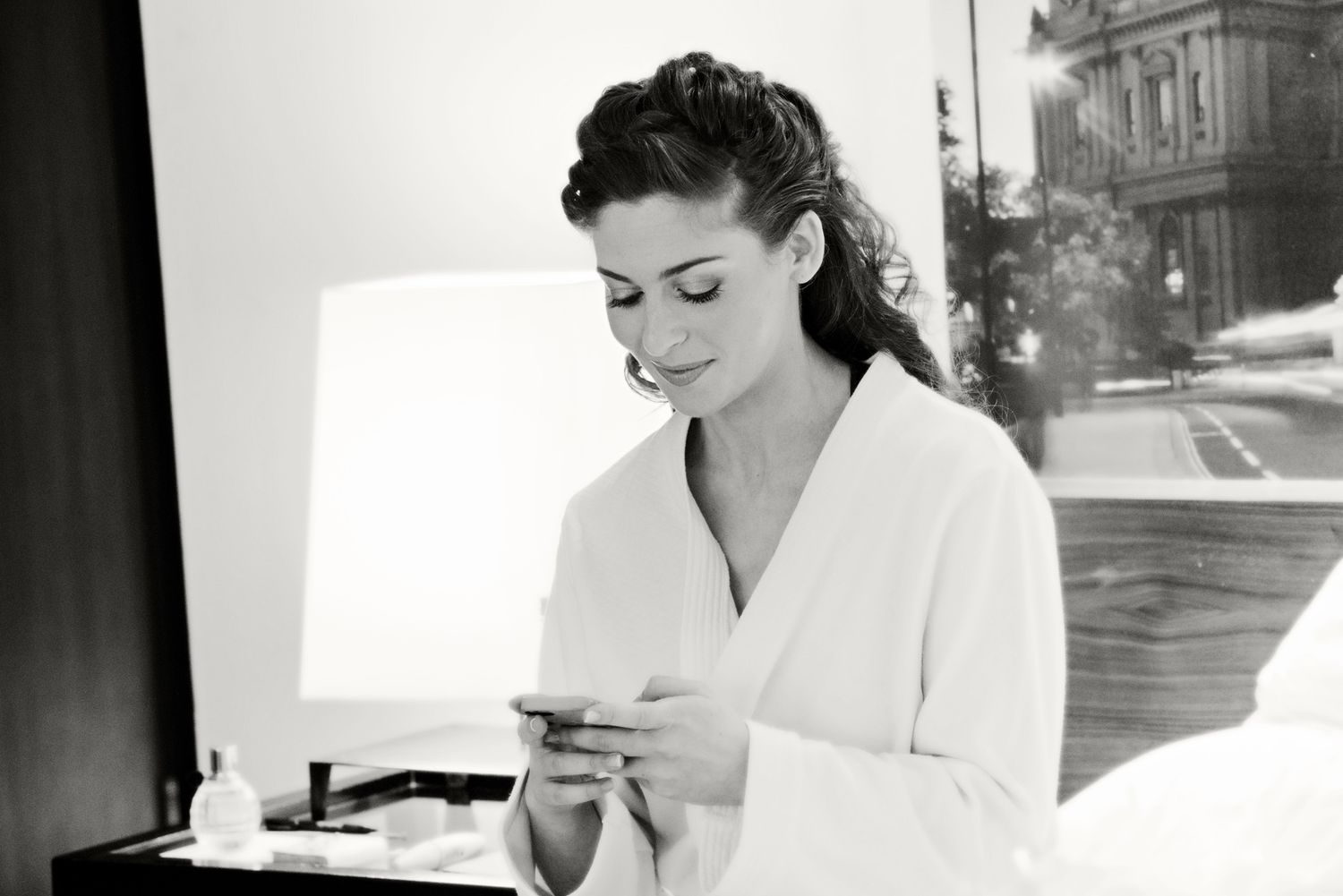 A woman in a white bathrobe smiles while using her smartphone in a bright, elegant hotel room.