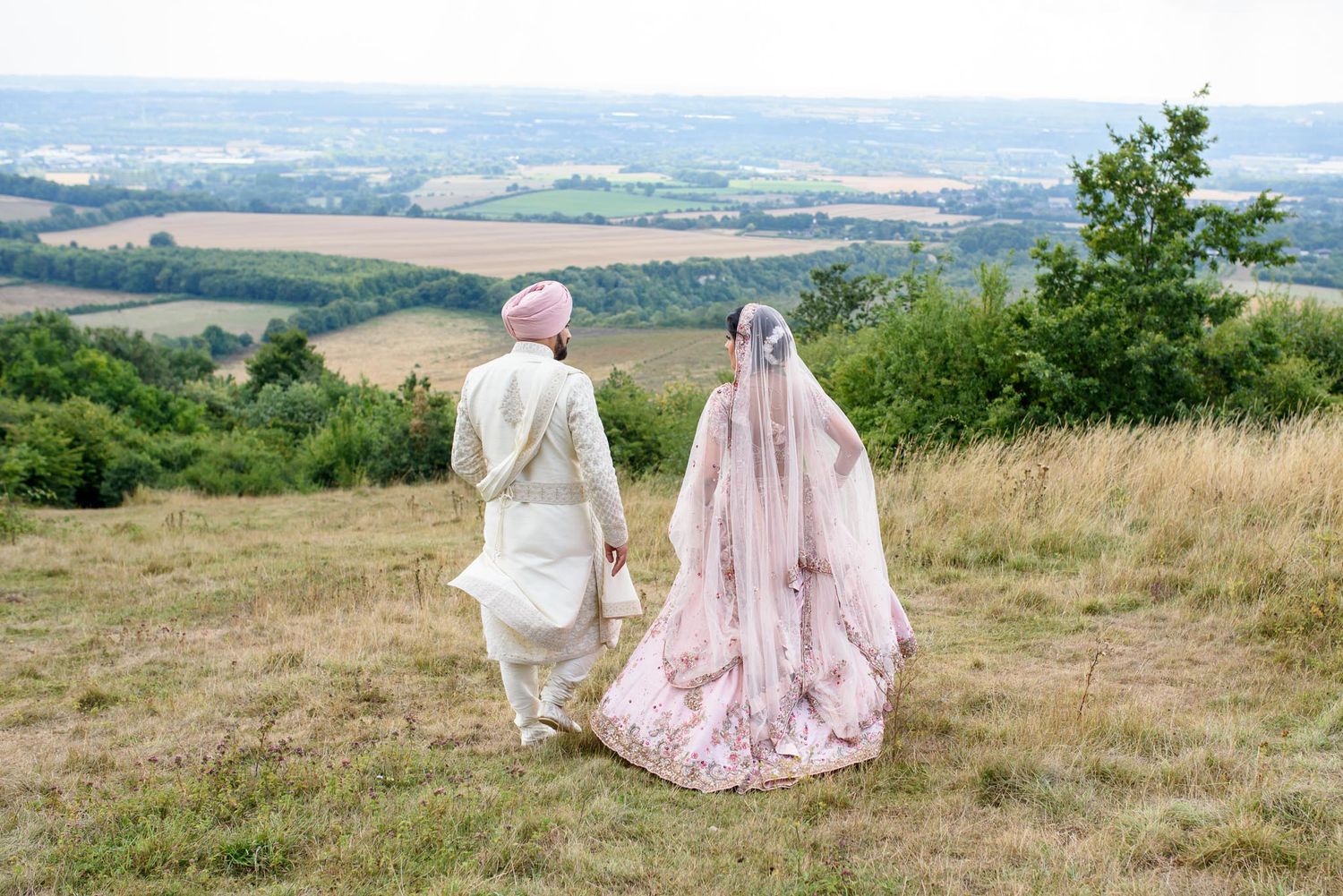 South Asian bride and groom in traditional attire holding hands overlooking a scenic countryside landscape.