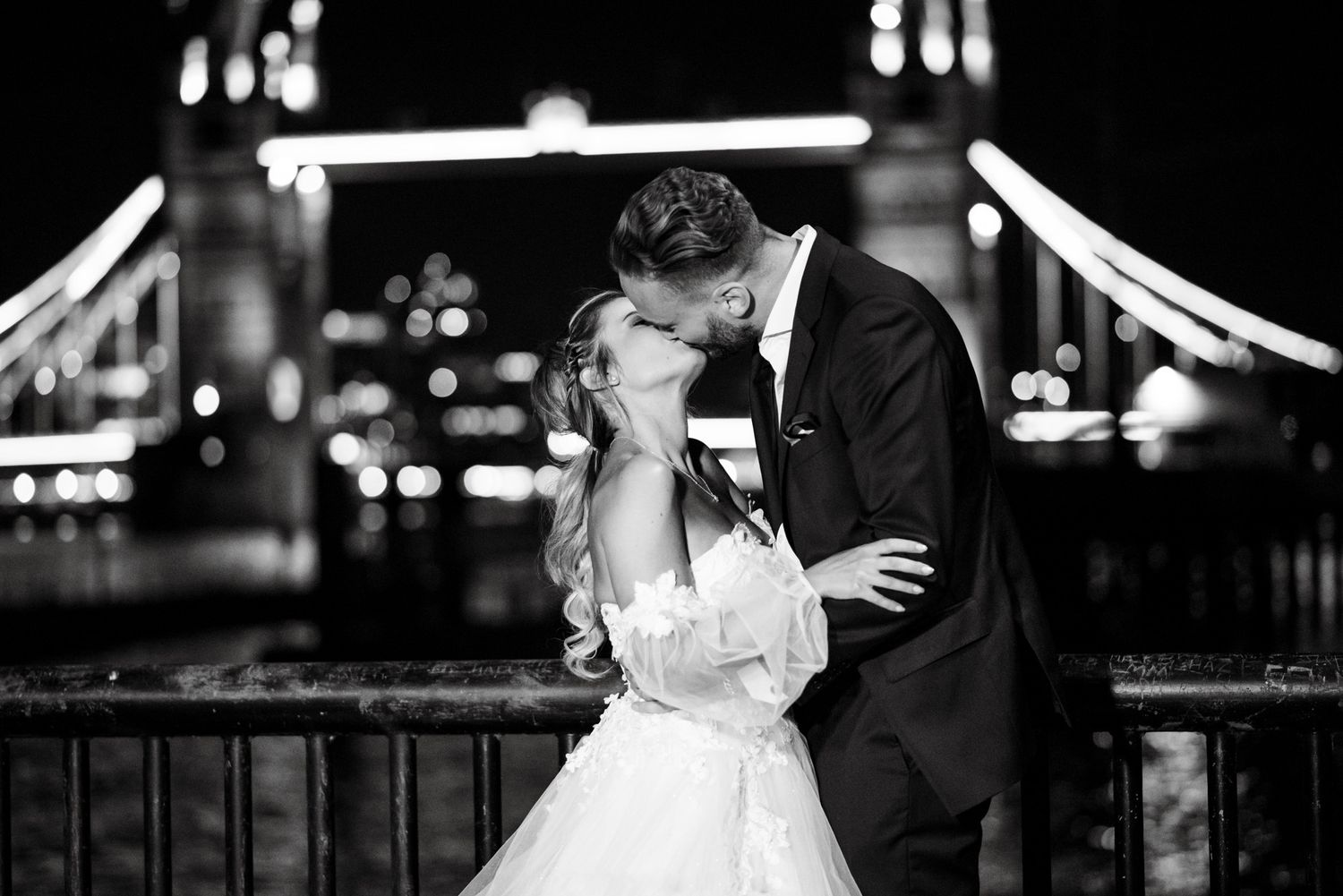 Black and white photo of a bride and groom sharing a kiss in front of Tower Bridge, London, at night.