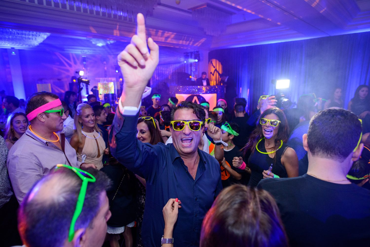 Energetic crowd dances and celebrates at a nightclub with blue and purple lighting effects and glowing accessories.