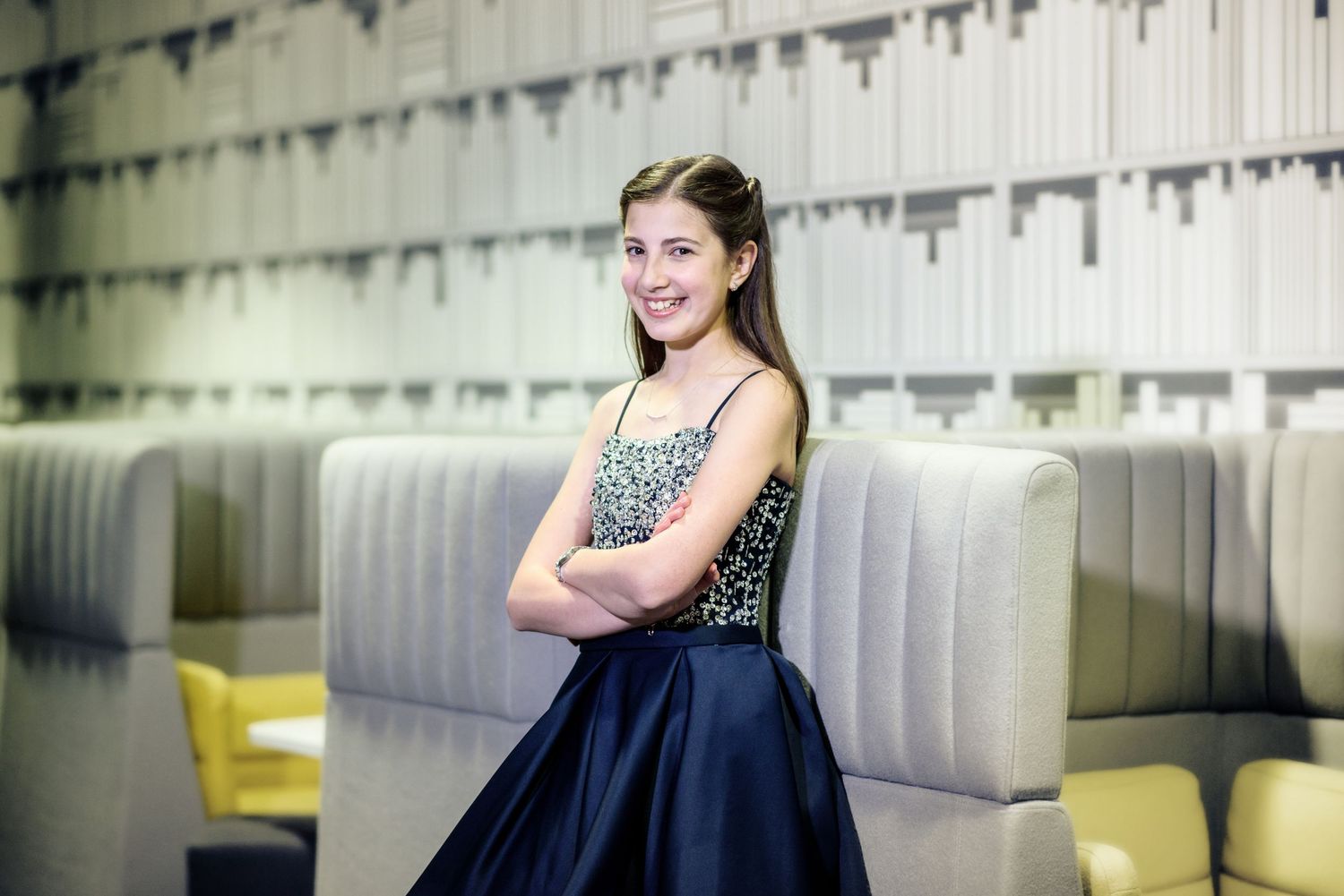 A person in a sleeveless sparkly top and navy blue skirt poses with crossed arms in front of a modern wall.