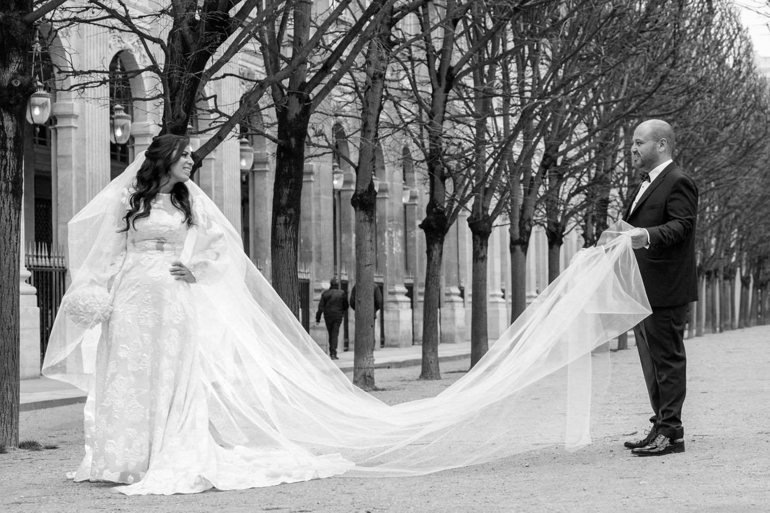 A long flowing wedding dress train billows in the wind along a tree-lined pathway in black and white.