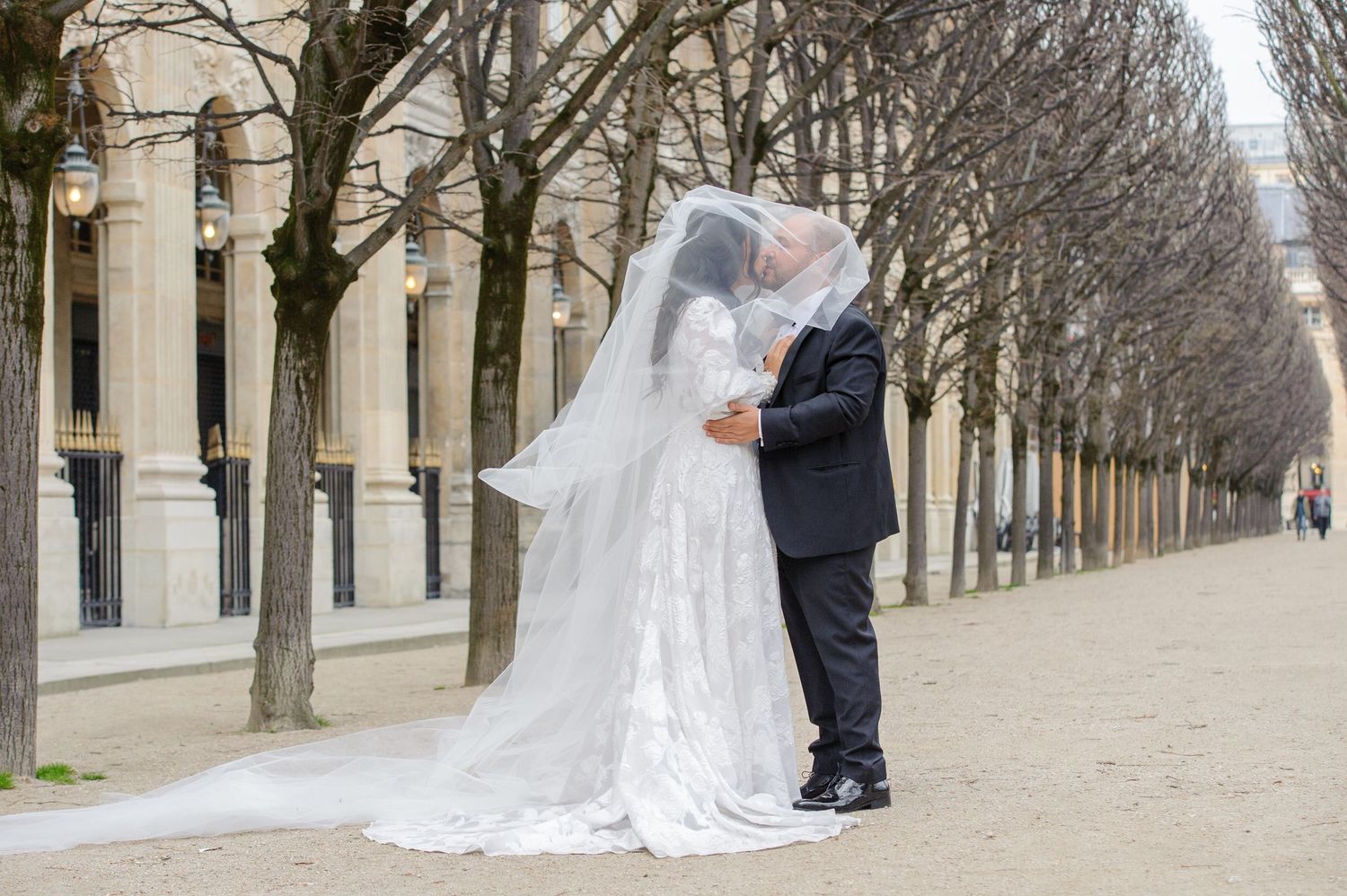 A romantic wedding couple walks along a tree-lined path in Paris, with the bride's veil flowing elegantly behind them.