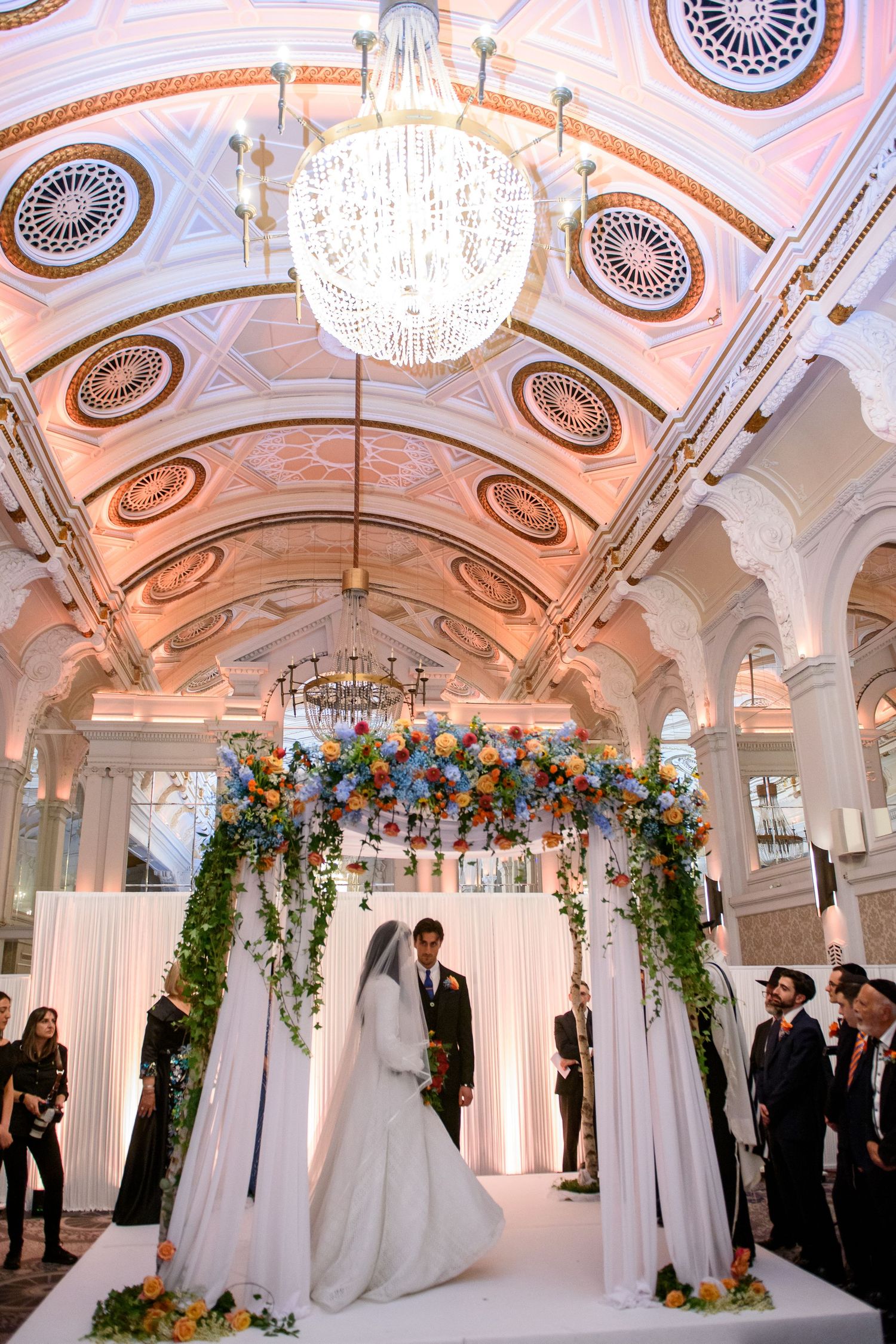 Elegant wedding ceremony setup in a grand ballroom with ornate ceilings, crystal chandeliers and floral arch decorations.