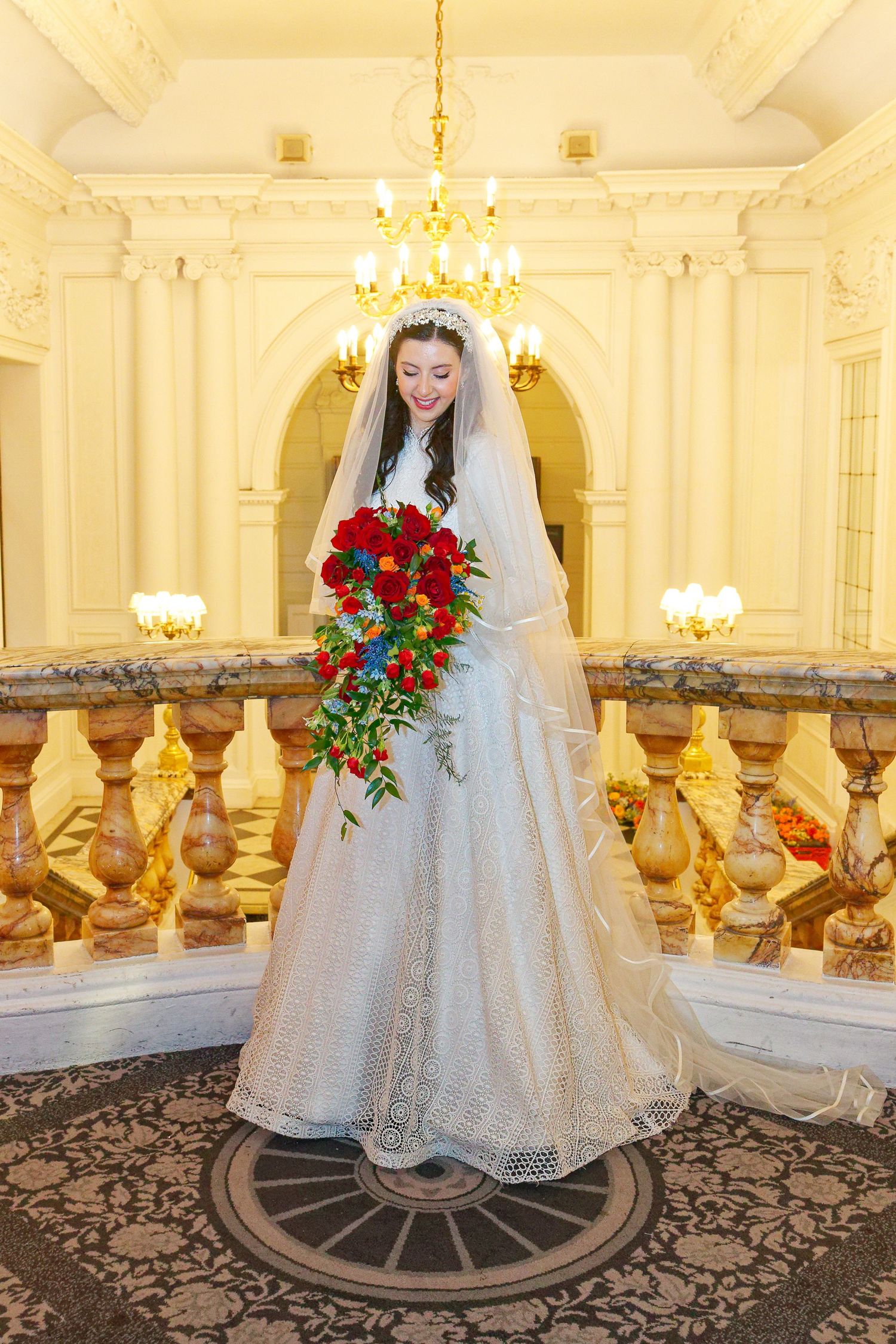 Elegant white lace wedding dress with red rose bouquet displayed in ornate ballroom with crystal chandeliers and patterned carpet.