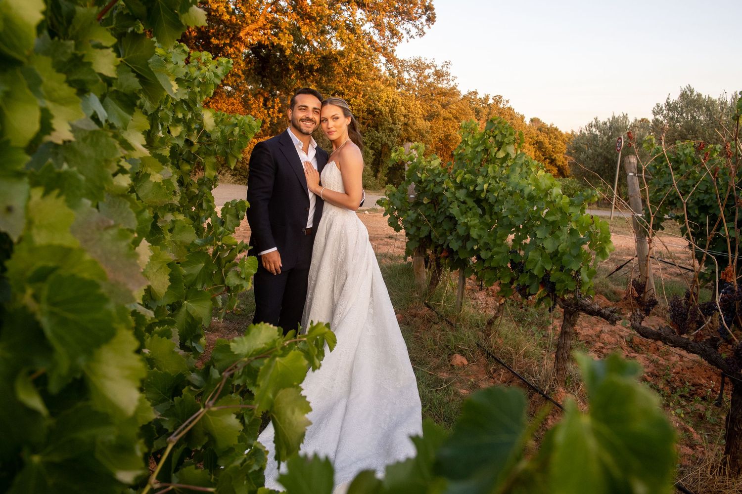 Couple embraces in a scenic vineyard during golden autumn sunset with colorful fall foliage in background.