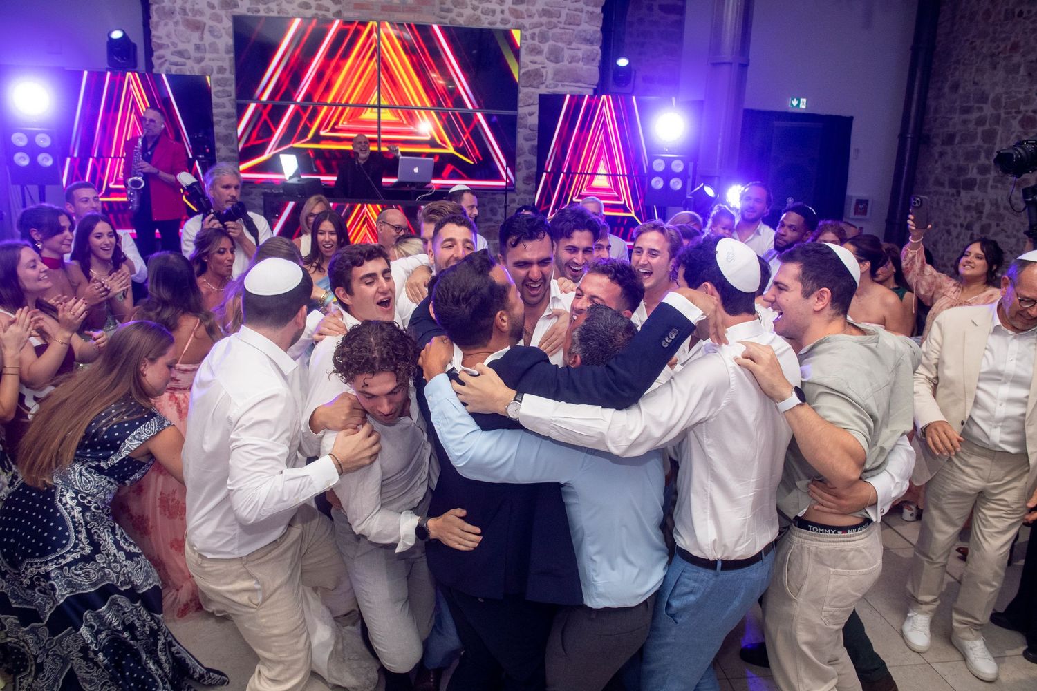 A large crowd dances and celebrates at a wedding reception with colorful neon triangles and purple lighting in the background.