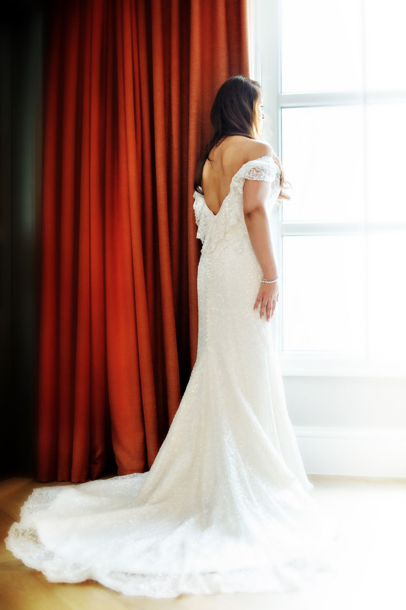 A wedding dress with lace details and long train shown from multiple angles against red curtains and natural light.