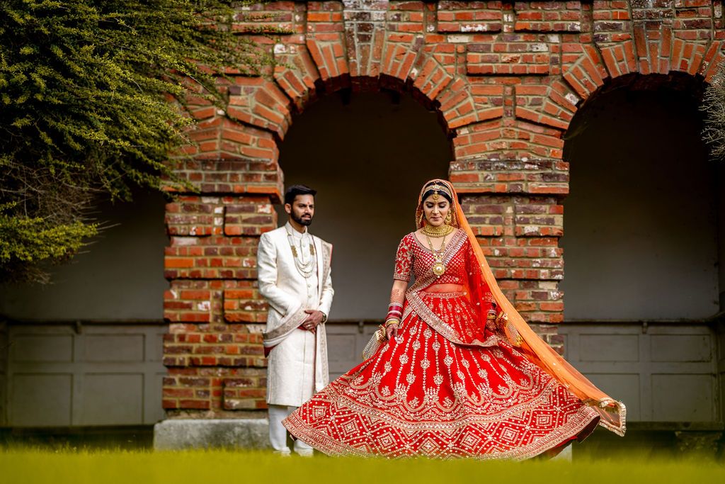 Indian bride in red lehenga and groom in white sherwani posing before brick arches at traditional wedding.