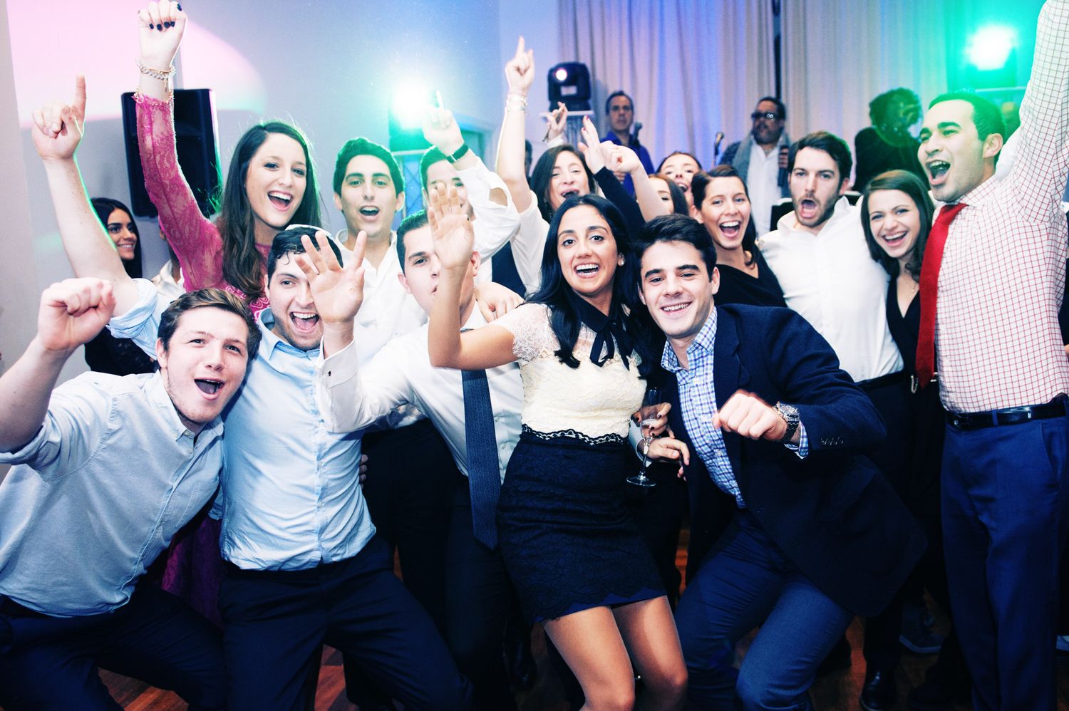 Energetic crowd of party guests dancing and celebrating with raised arms on a dance floor with blue lighting.