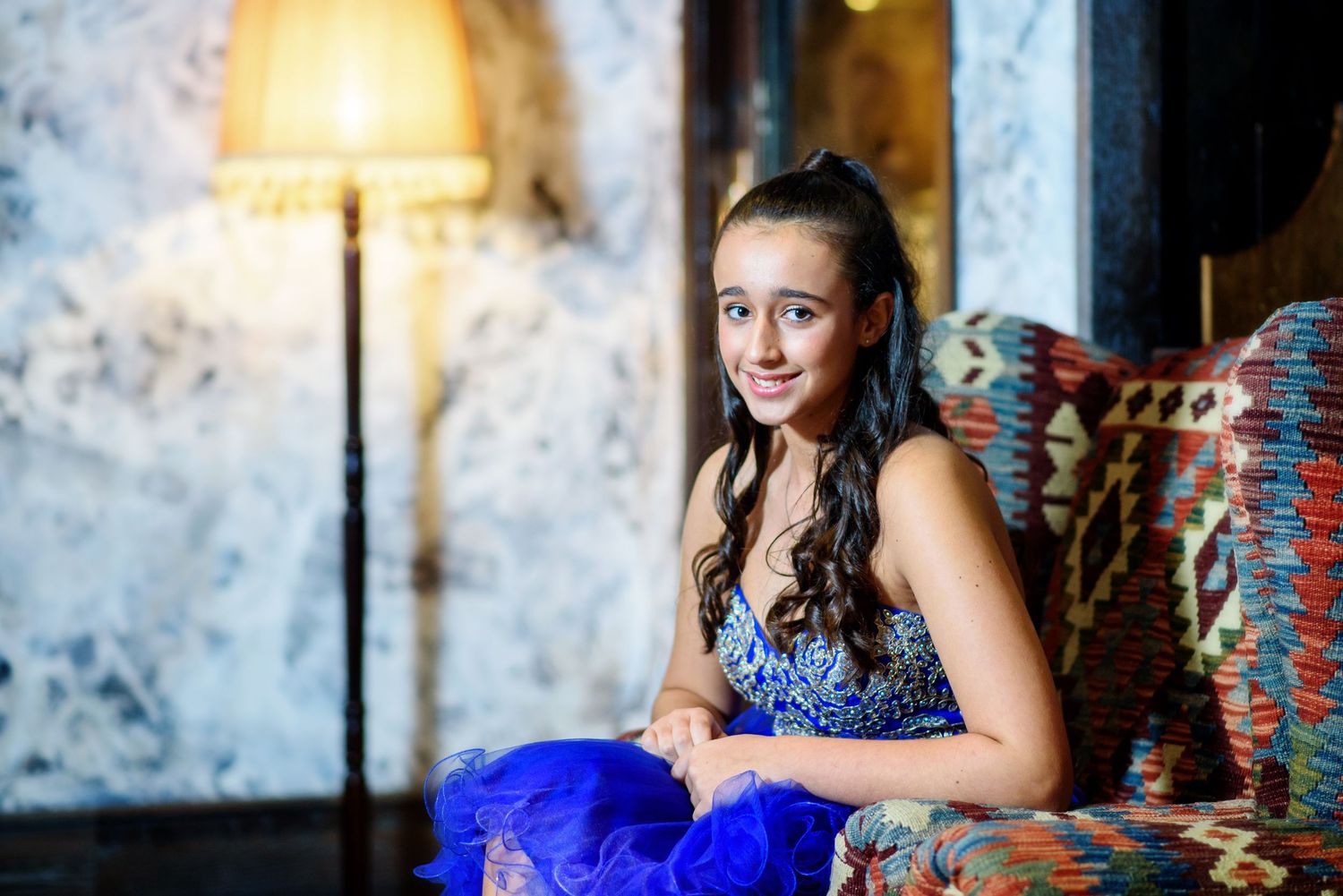 A person in a royal blue formal gown poses elegantly against a textured wall background.