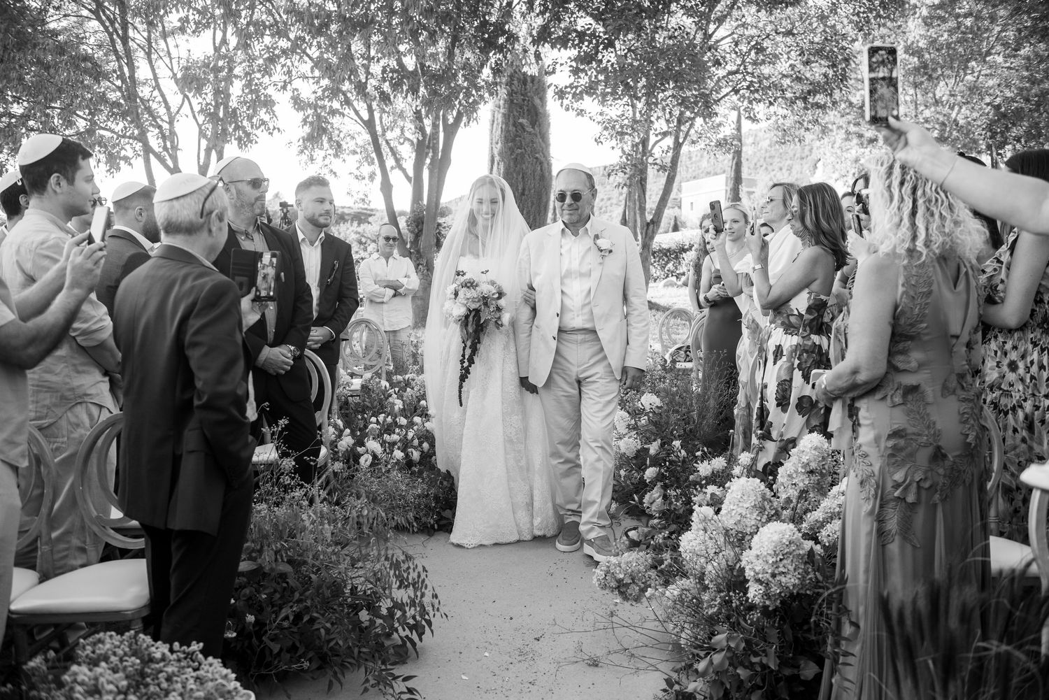A black and white wedding ceremony photo showing guests gathered outdoors among trees and garden scenery.