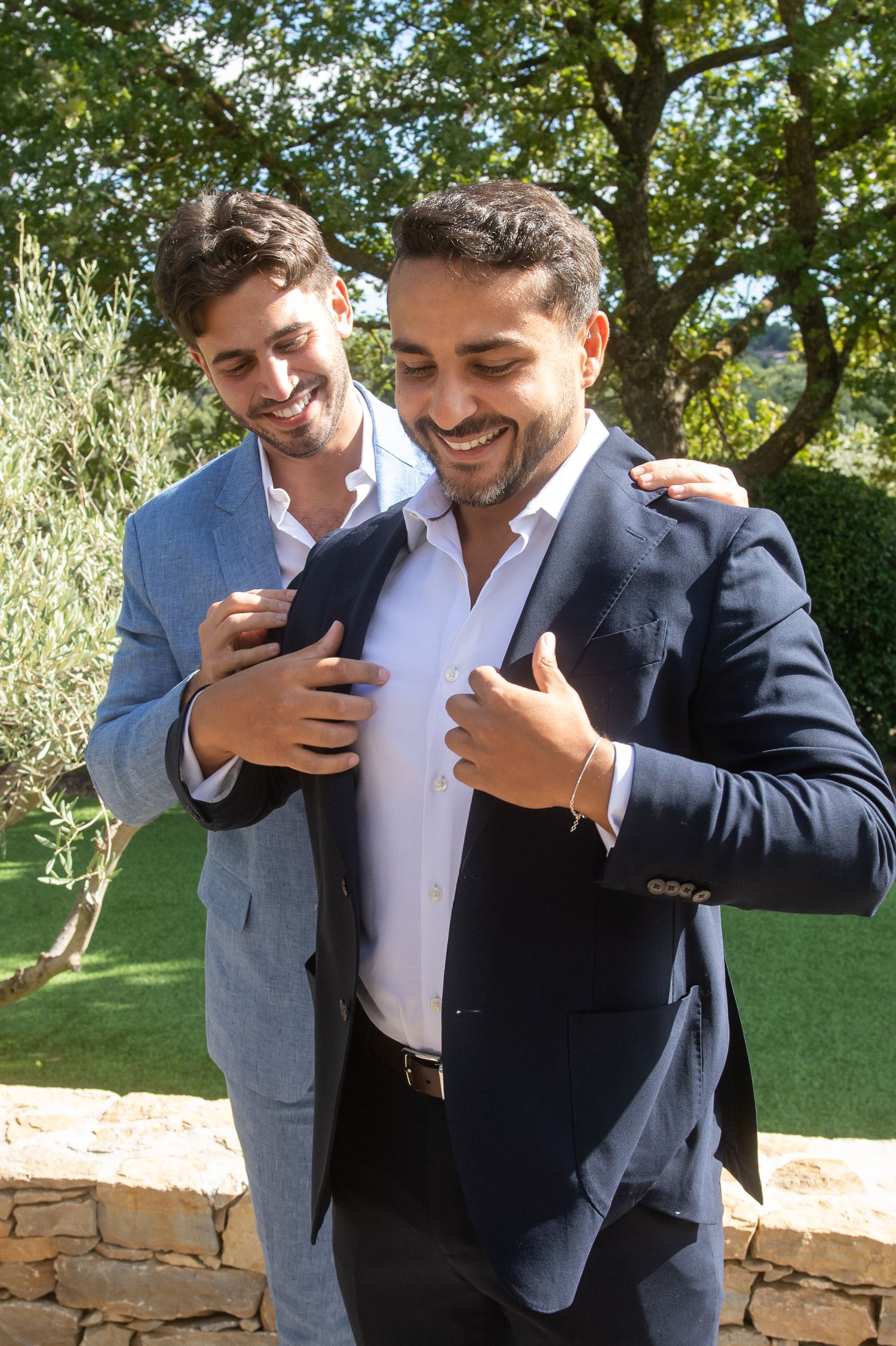 Two men in formal attire share a lighthearted moment while looking at a phone screen outdoors.