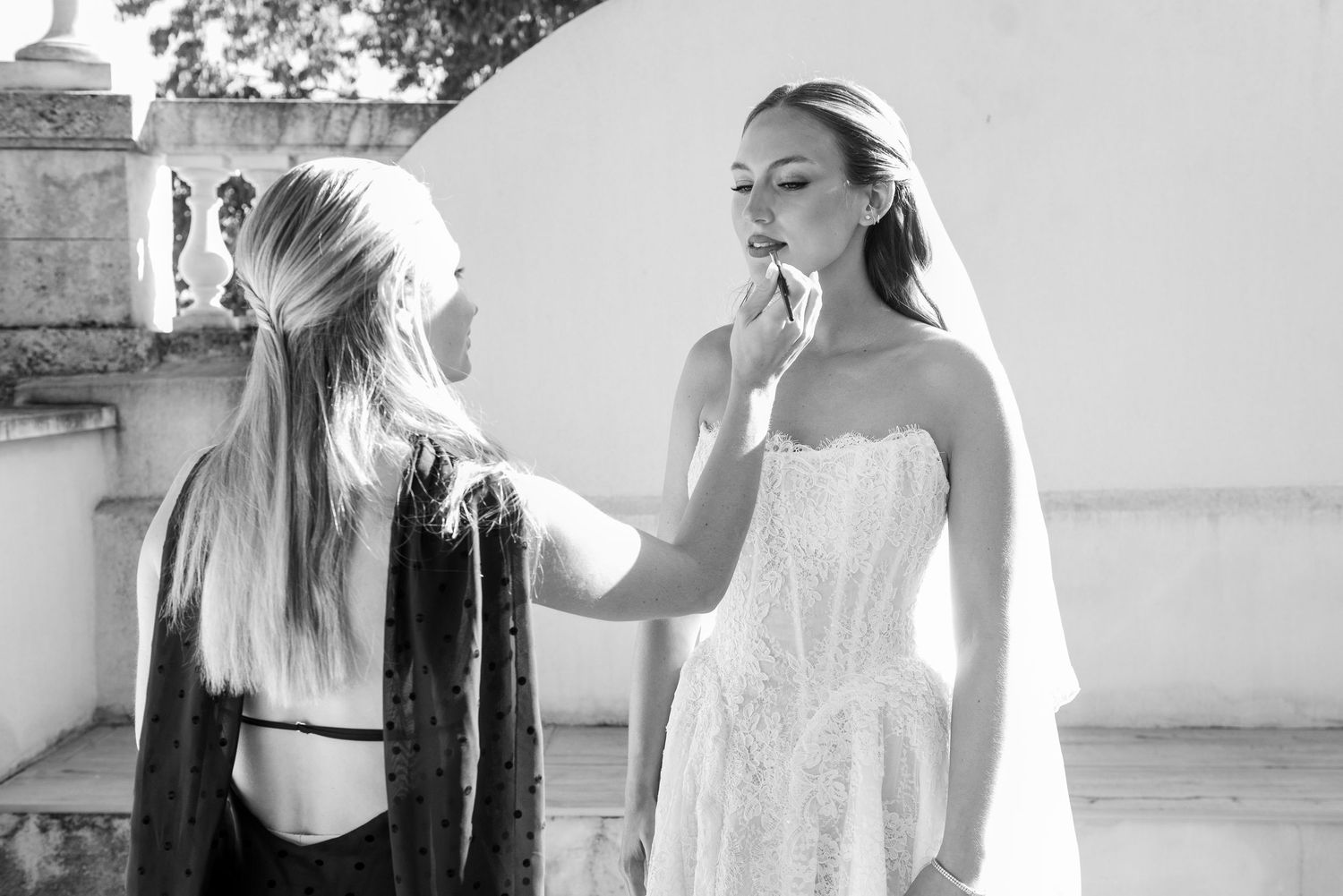 Black and white photo of a makeup artist applying lipstick to a bride in a strapless lace wedding gown outdoors.