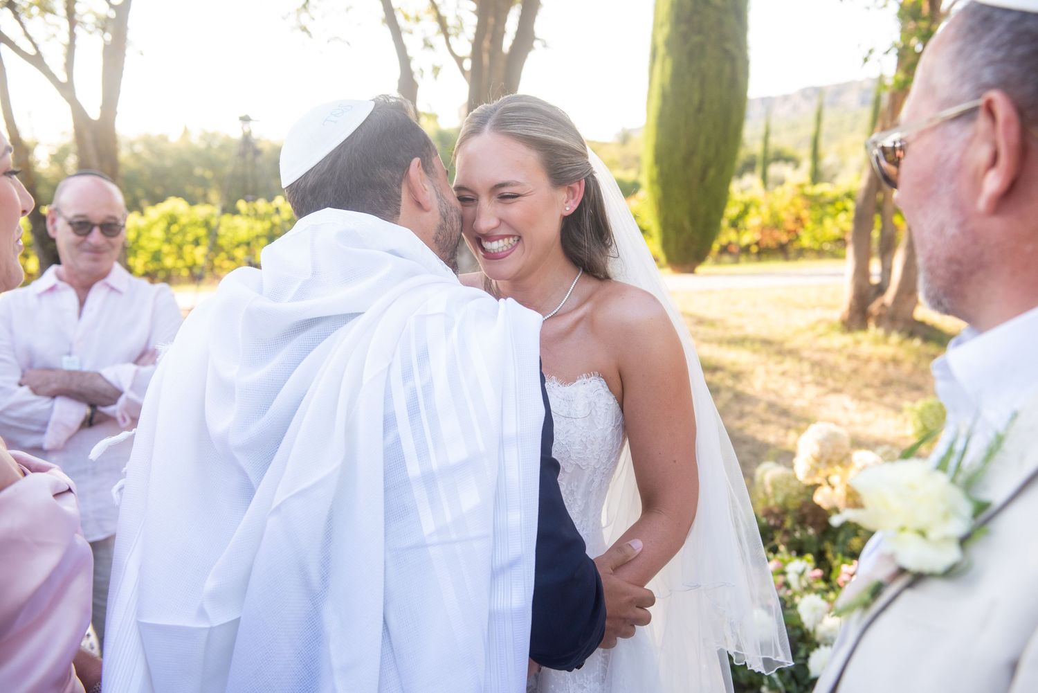 Bride smiling joyfully during an outdoor wedding ceremony, wearing a white dress while greeting a guest in white robes.