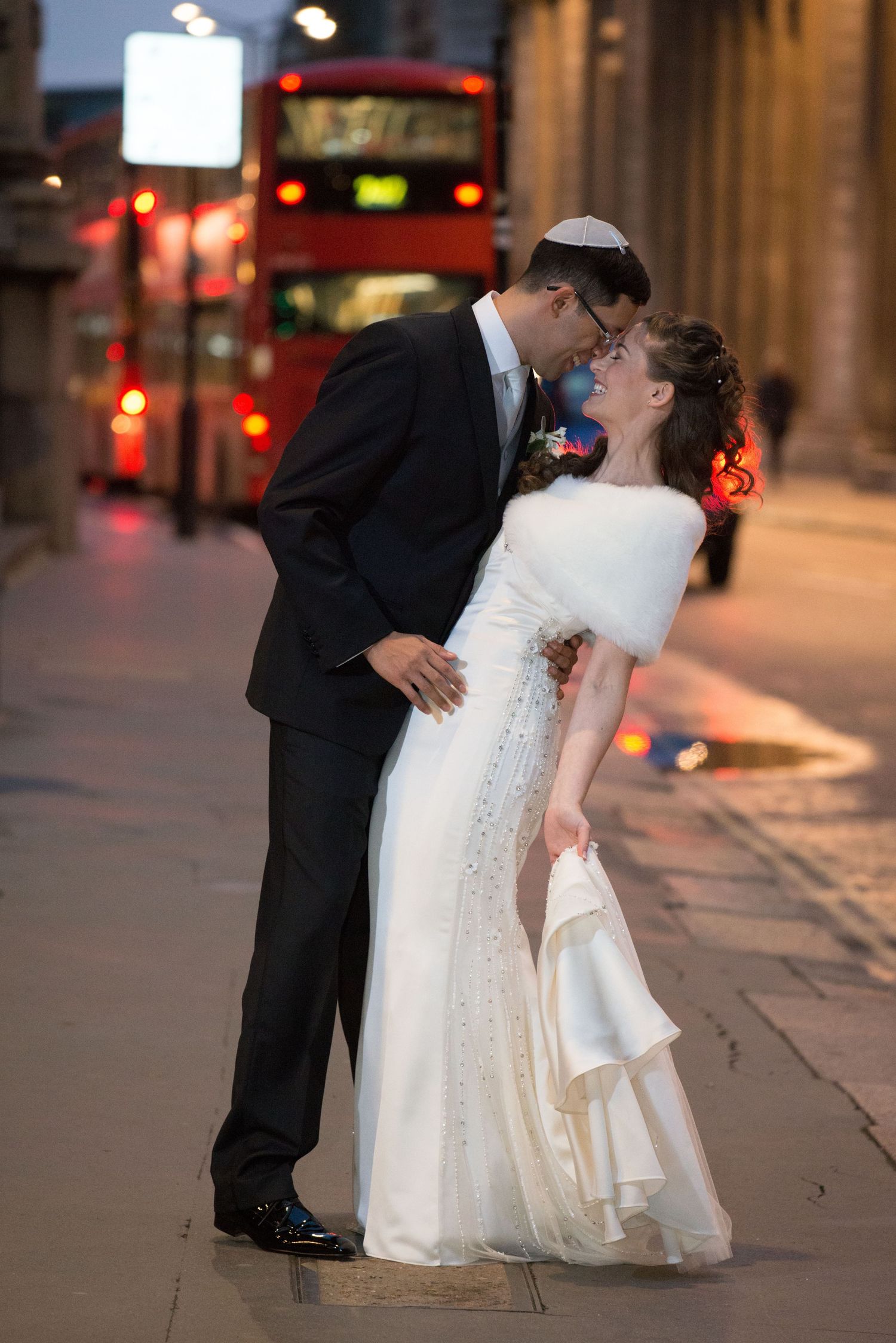 Newlywed couple sharing a romantic kiss on a London street with a red double-decker bus in the background.