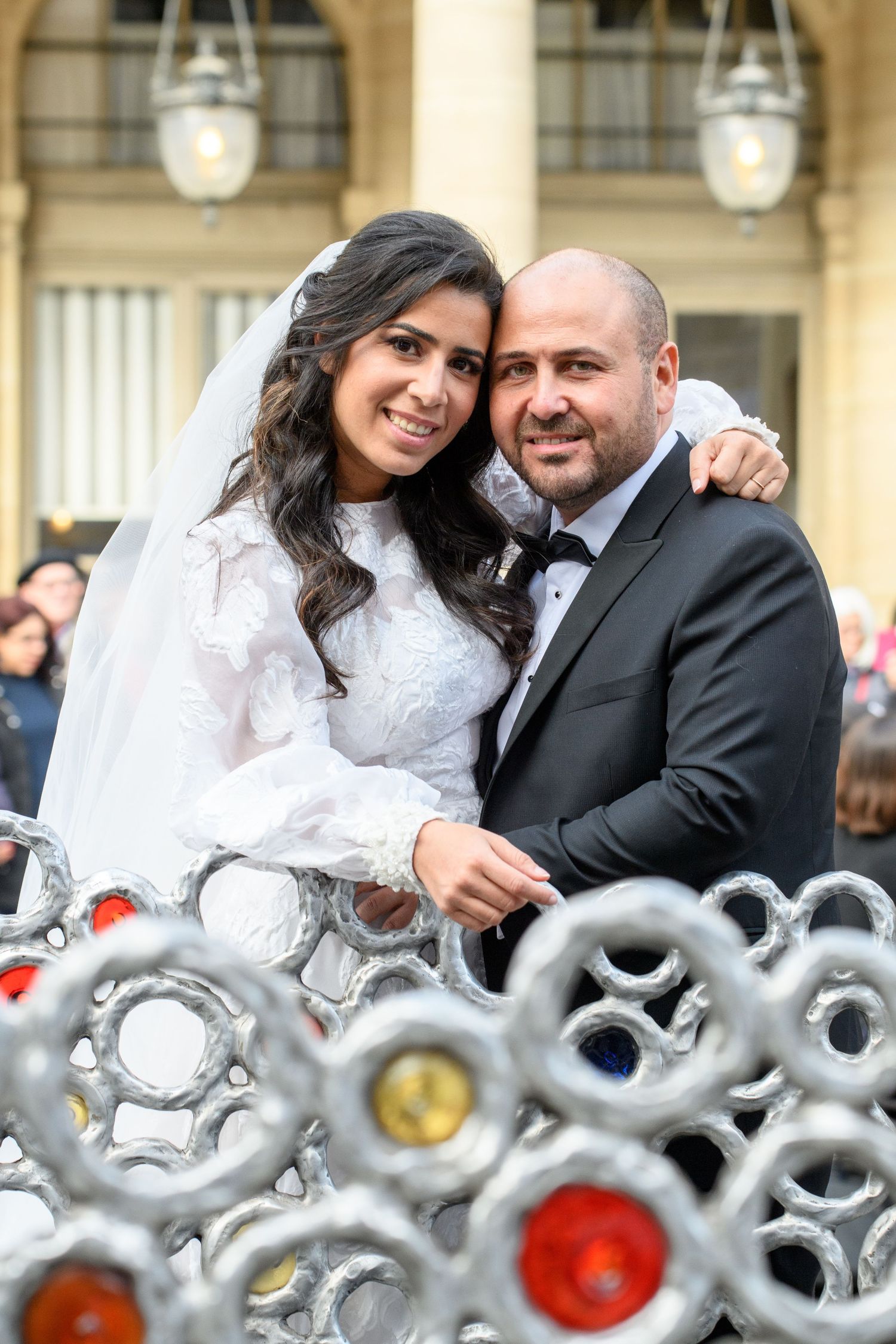 A happy wedding couple poses together behind an ornate iron fence with a grand building facade in the background.