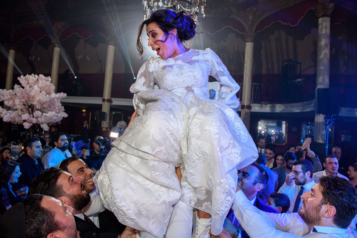A bride in a white gown is lifted by guests during a reception under blue lighting.