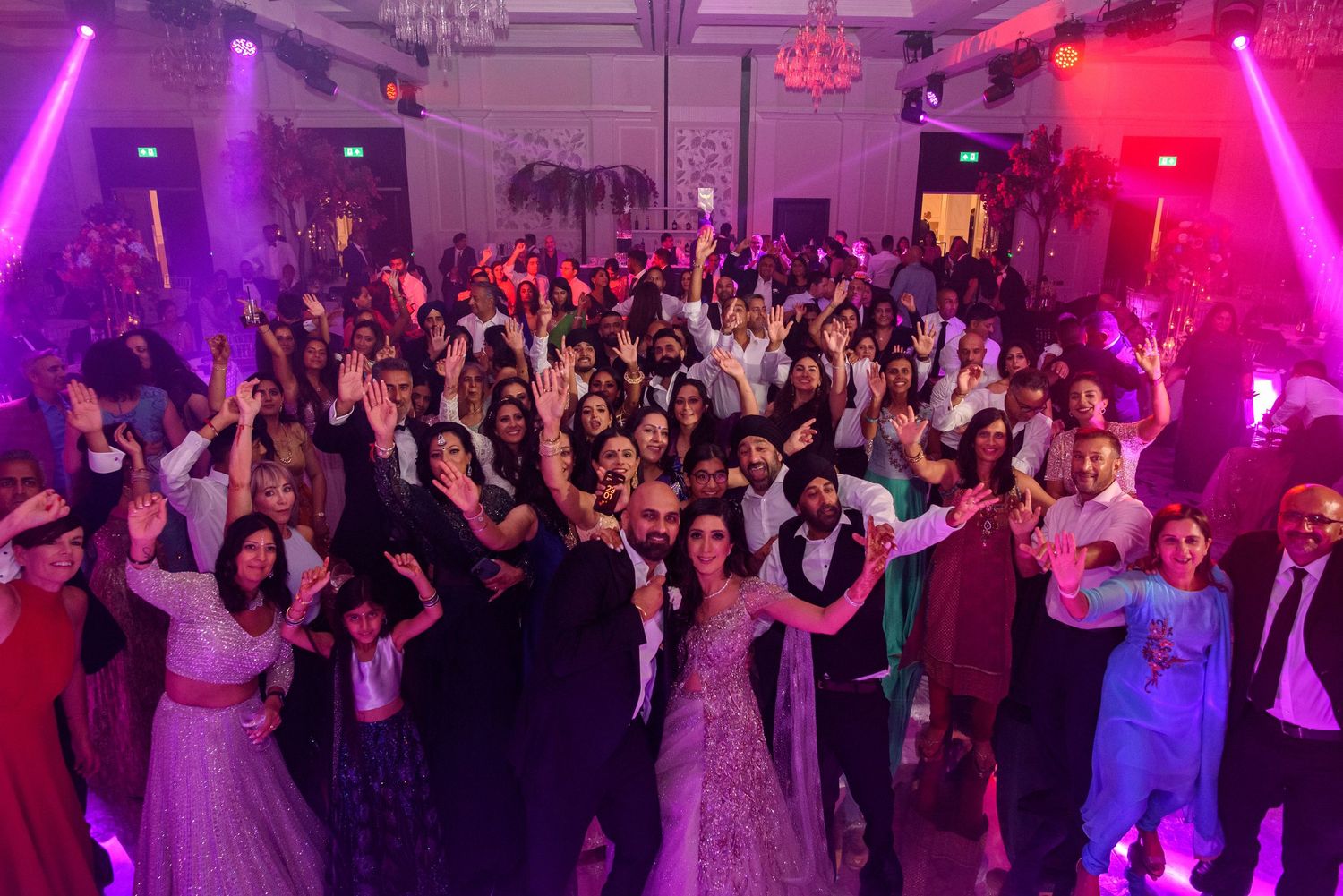 Wedding reception guests dancing and celebrating on a dance floor illuminated by vibrant pink and purple lighting.