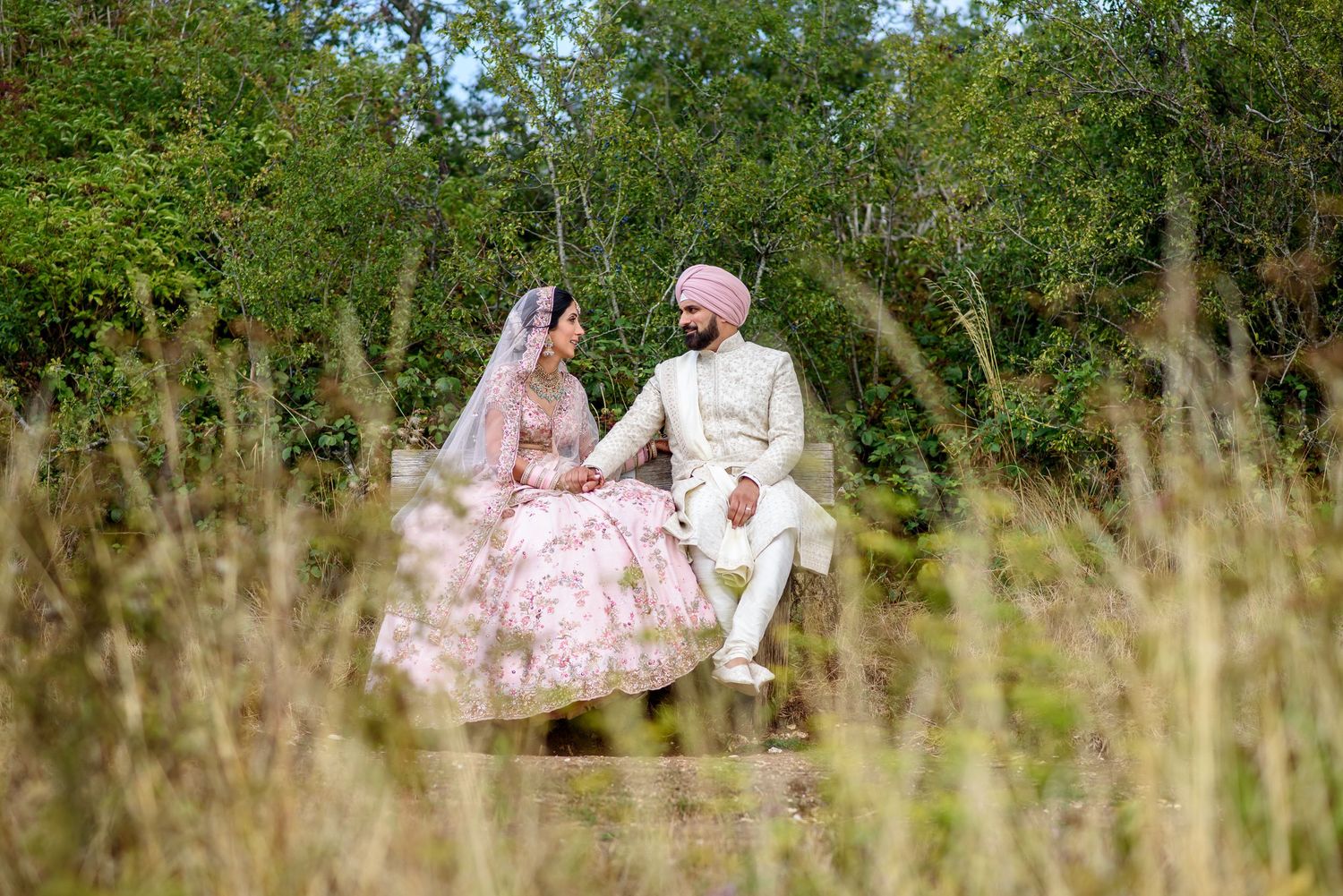 A couple in traditional Indian wedding attire poses together in a lush green garden setting.