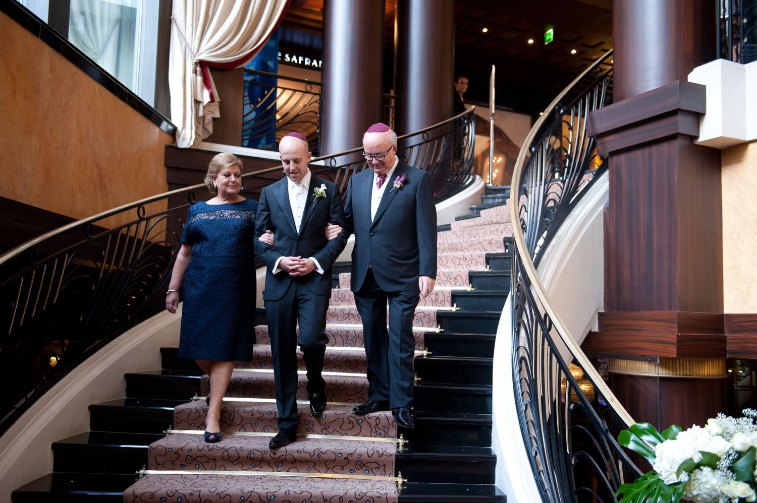 Three people walk down an elegant curved staircase with wooden columns and railings in a formal setting.