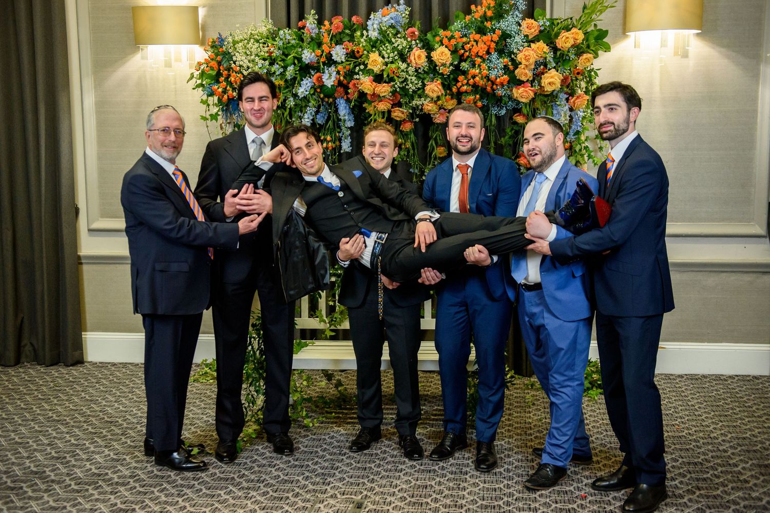 A group of wedding guests in blue and black suits gather around colorful floral arrangements at an elegant venue.
