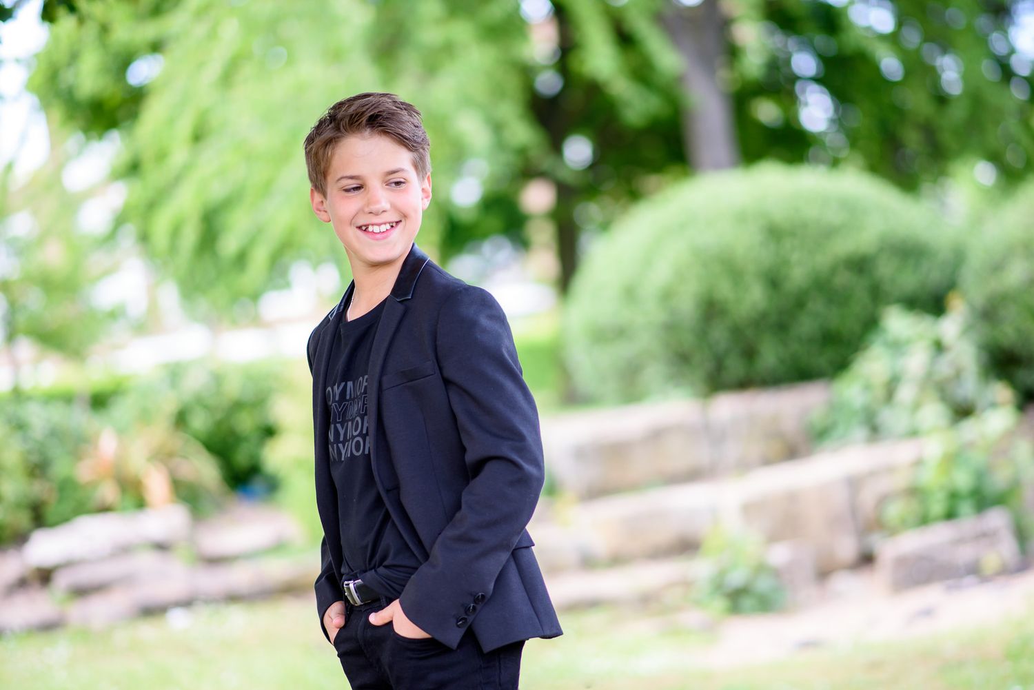 A child wearing a navy blue blazer poses outdoors against a blurred green garden background.
