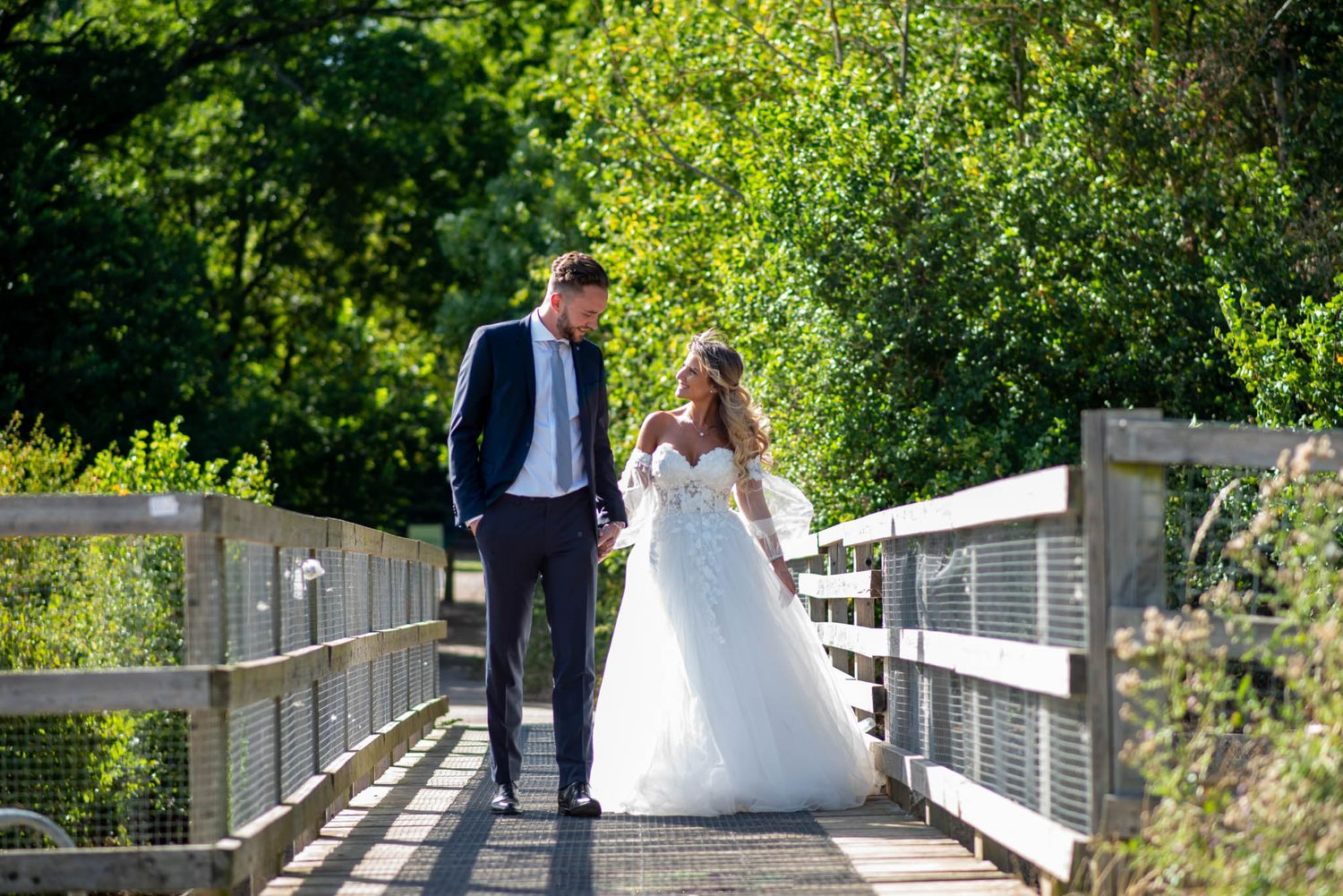 A bride and groom walk hand-in-hand on a wooden bridge surrounded by lush green trees on their wedding day.