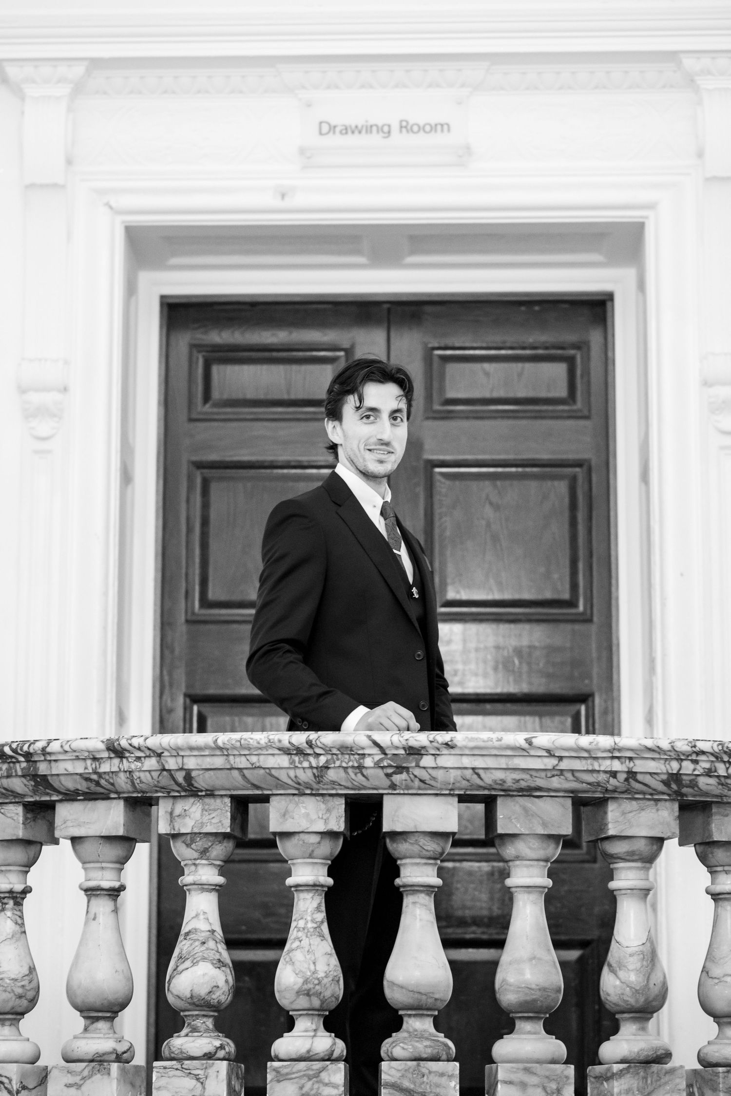A person in formal attire stands behind a white stone balustrade with ornate balusters outside a building.