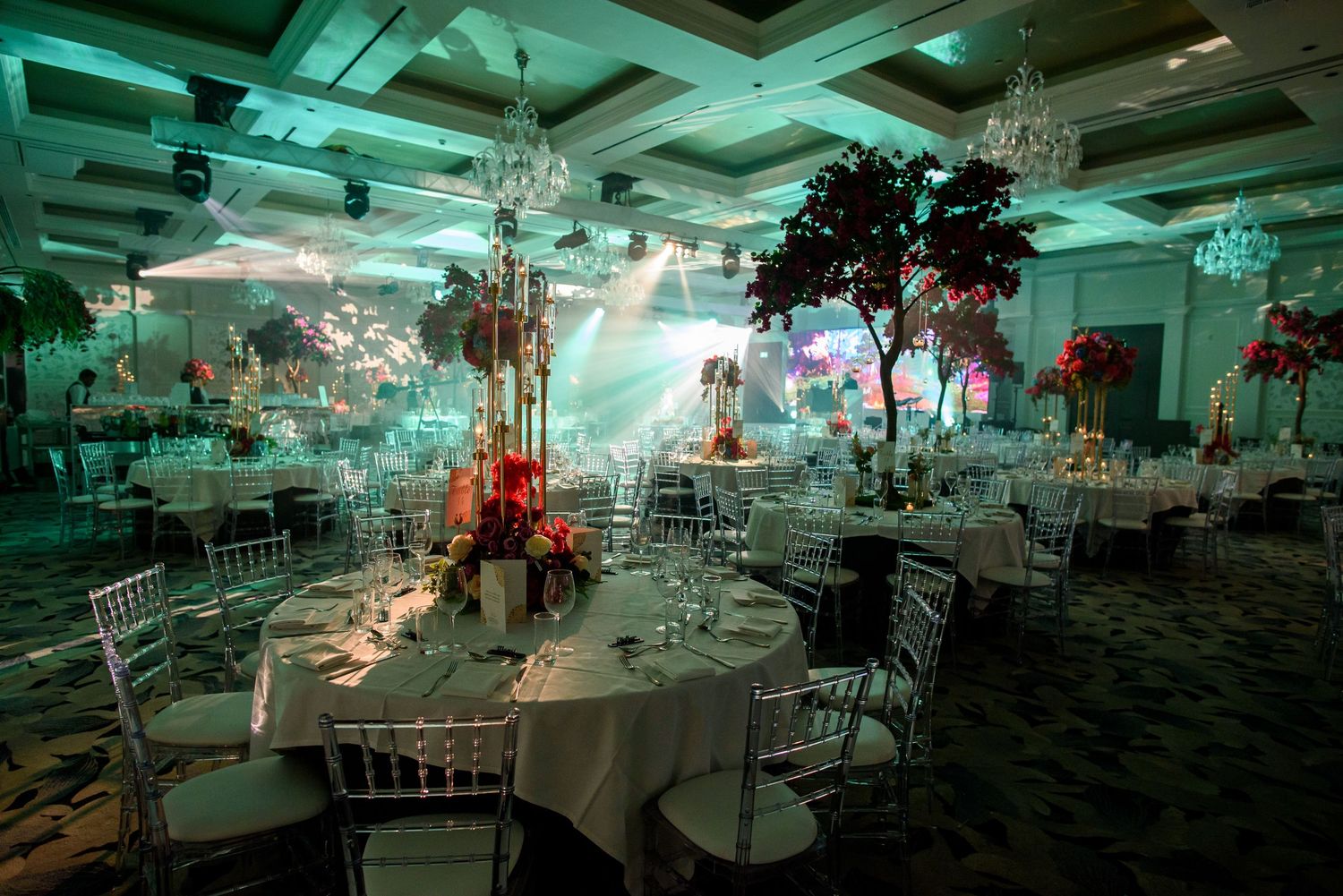 Elegant banquet hall decorated with crystal chandeliers, white tablecloths and soft teal lighting for a formal event.