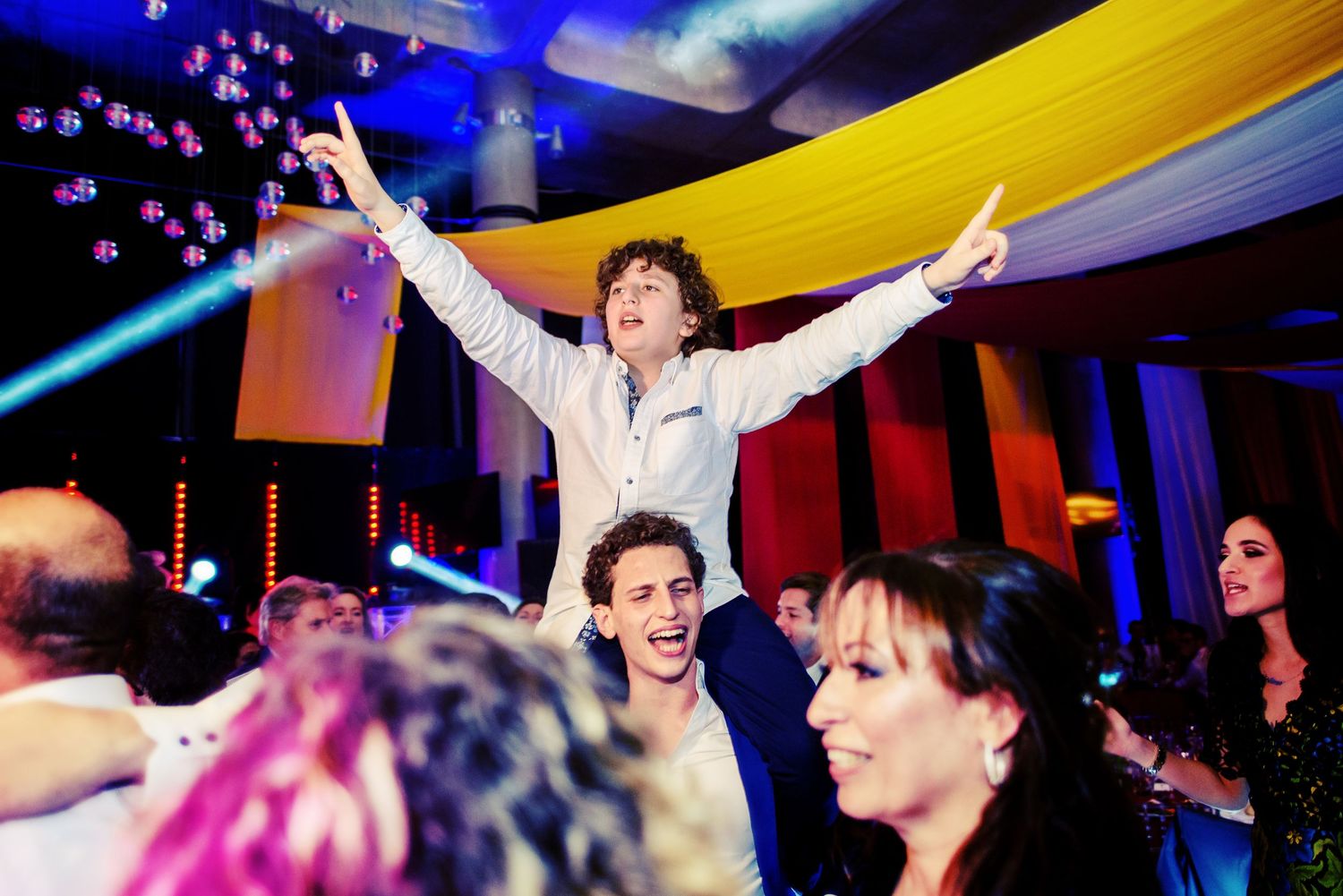A crowd of people dancing and celebrating at a nightclub with colorful lighting and fabric draping from the ceiling.