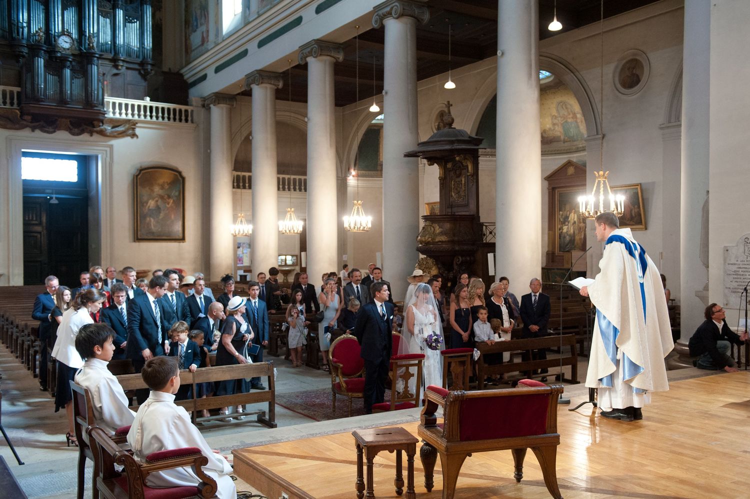 Wedding ceremony inside a grand church with guests seated in pews as a priest officiates the couple at the altar.