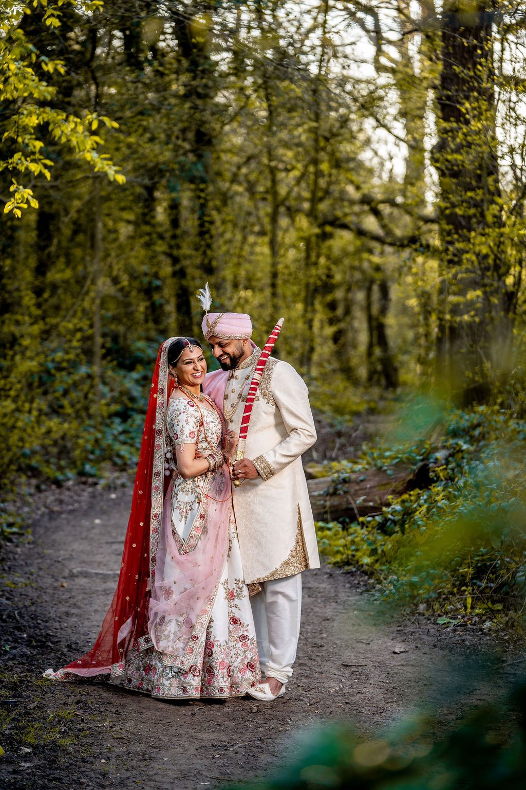 Indian bride and groom in traditional wedding attire stand together on a forest path surrounded by lush green trees.