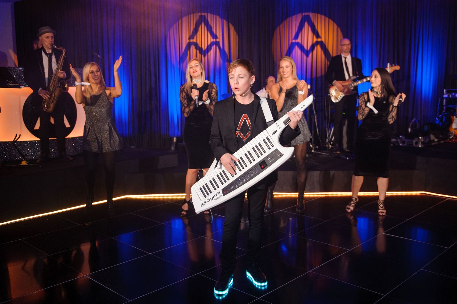 Musicians perform on stage with blue lighting and a keytar during an energetic concert performance.